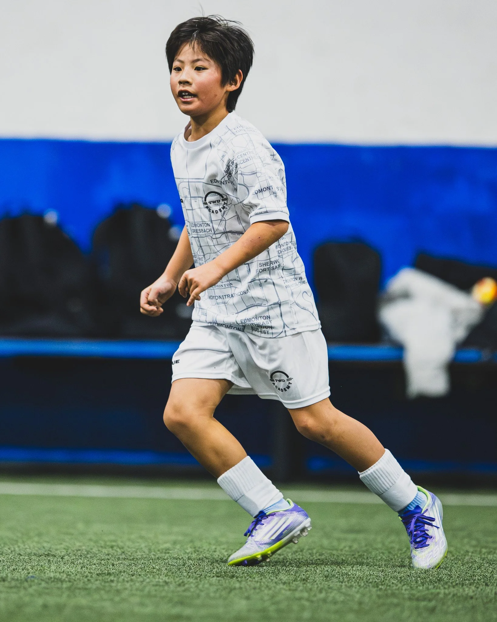 Young male soccer player running on a green indoor turf field wearing a white jersey with black city map print and matching shorts, purple and white cleats, and white socks.