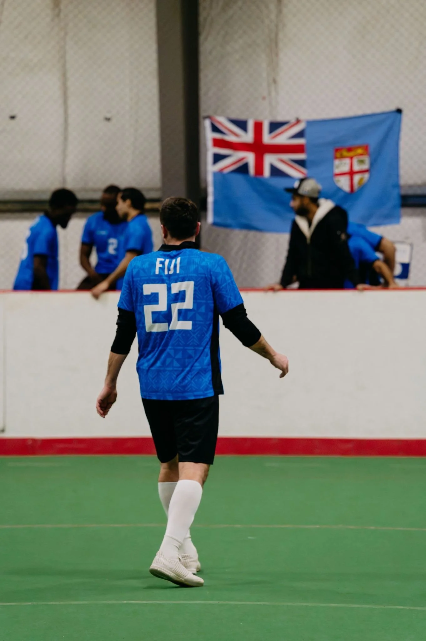 A soccer player wearing a blue jersey with number 22 and 'FIJI' on the back, running on an indoor field. In the background, a group of people are gathered near a wall with a Fiji flag.