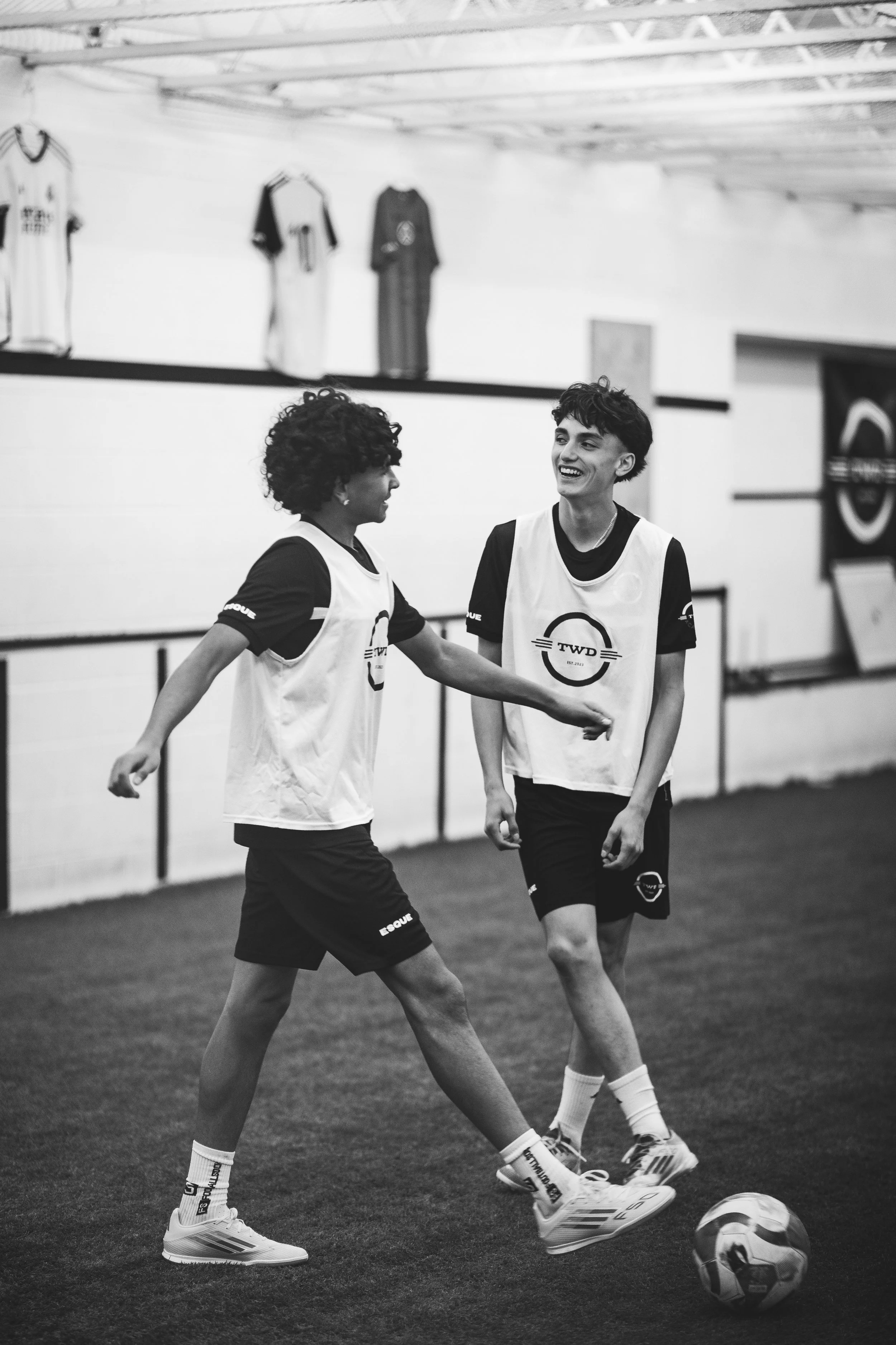 Two young women in sports jerseys and shorts playing soccer indoors. One is kicking the ball toward the other, who is smiling. Jerseys have a logo and the text "TWD."