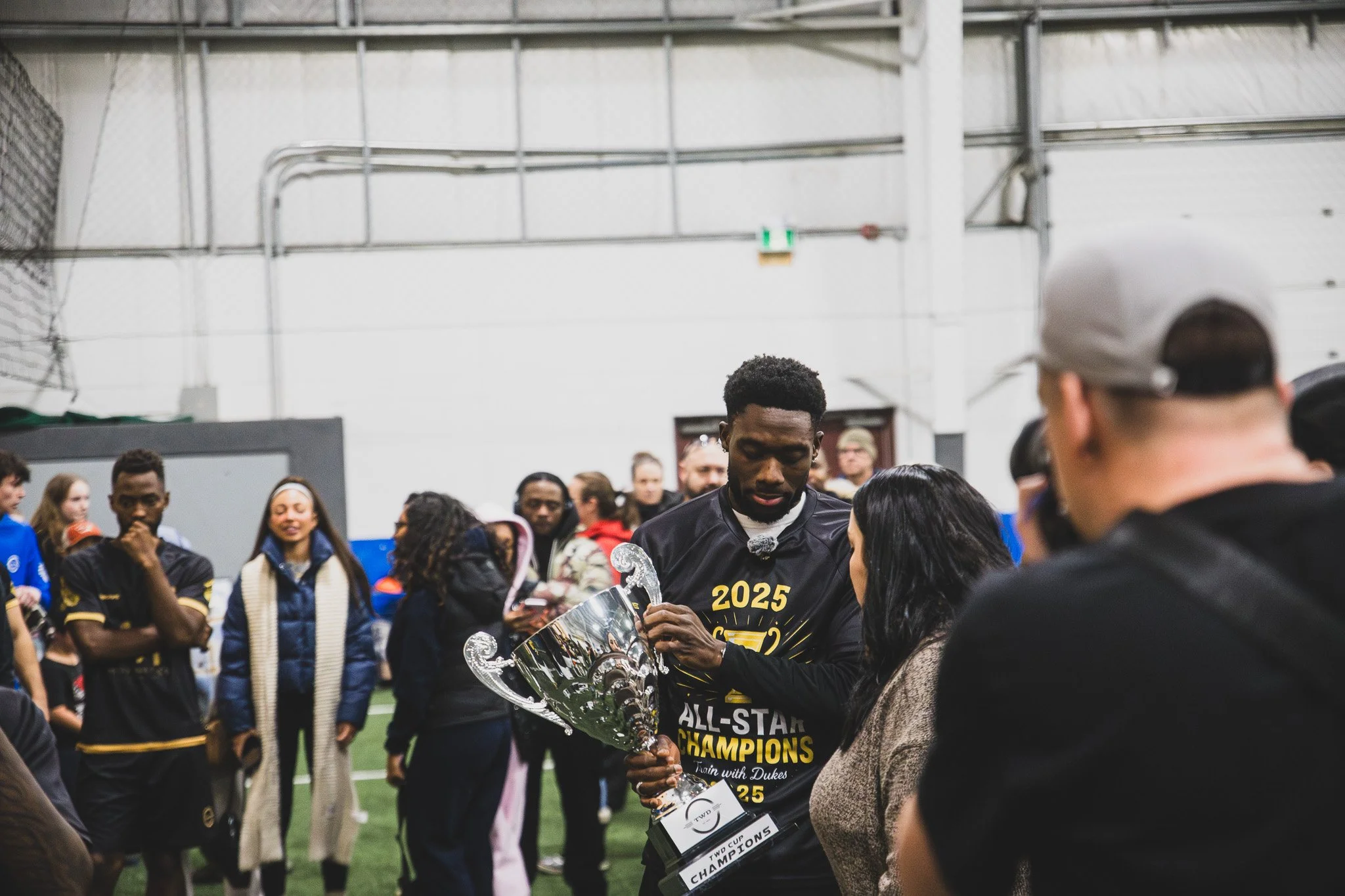 A man holding a trophy, surrounded by people, at an indoor sports event or celebration, with many onlookers and a high ceiling.