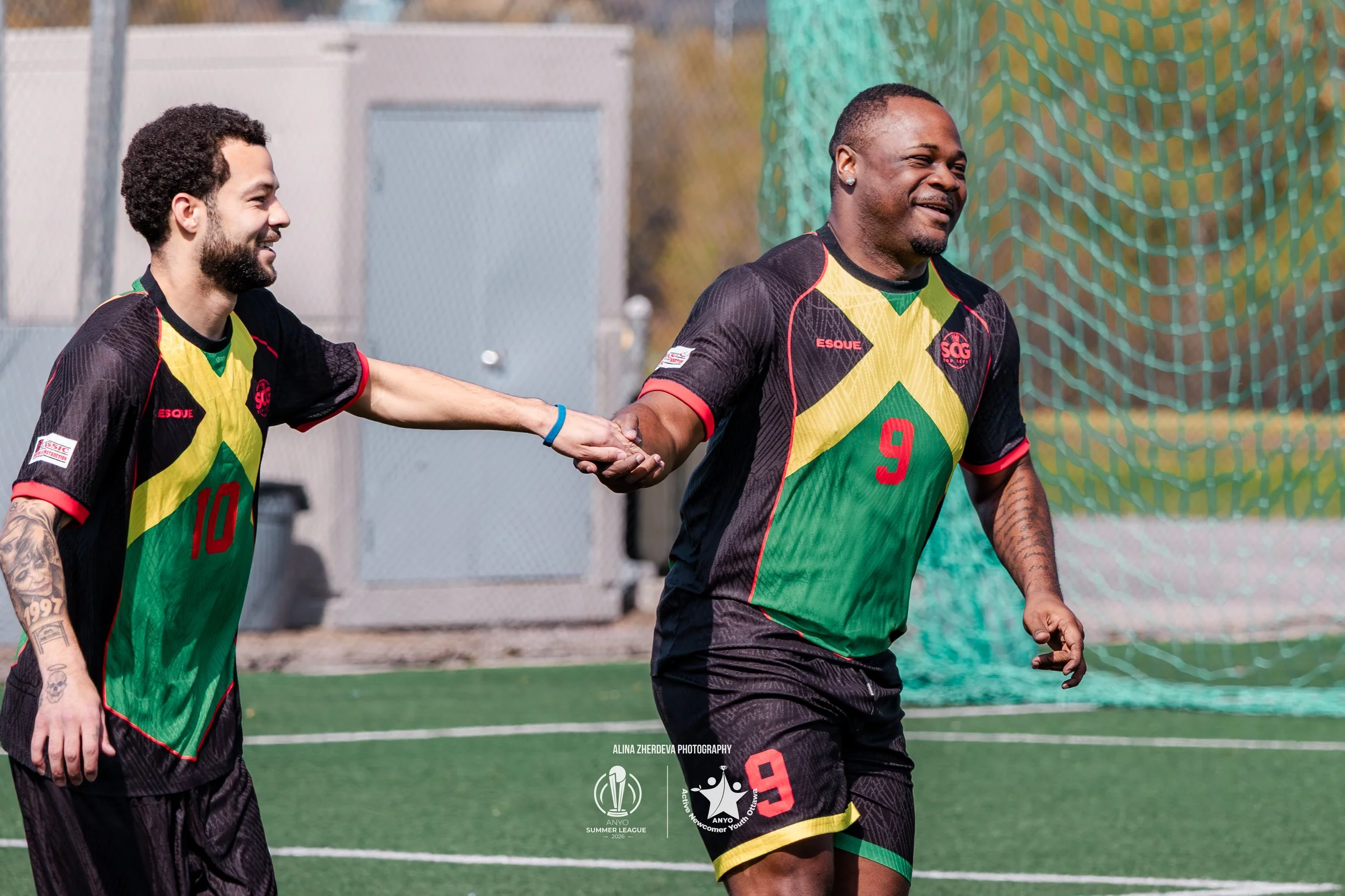 Two soccer players holding hands and smiling on a soccer field. Both are wearing black jerseys with yellow, green, and red accents. One player has the number 10, and the other has the number 9. There is a net and some trees in the background.
