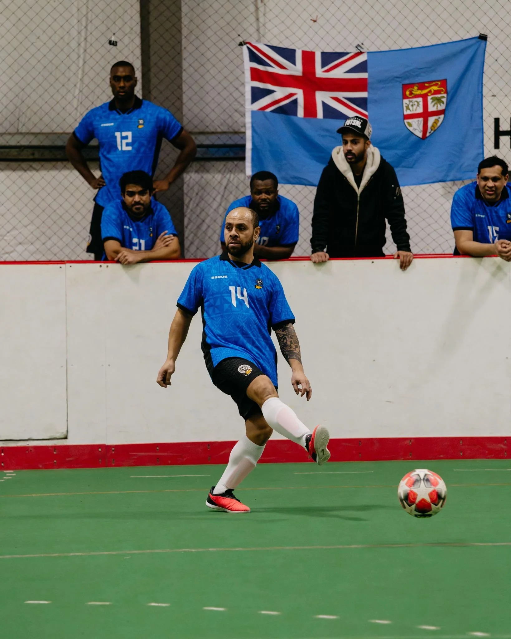 A soccer player in a blue jersey with the number 14 is kicking a soccer ball on an indoor field with a green surface. Behind him, five other players in blue jerseys are watching. There is a blue flag with the Union Jack and a crest hanging on a wall with a chain-link fence.