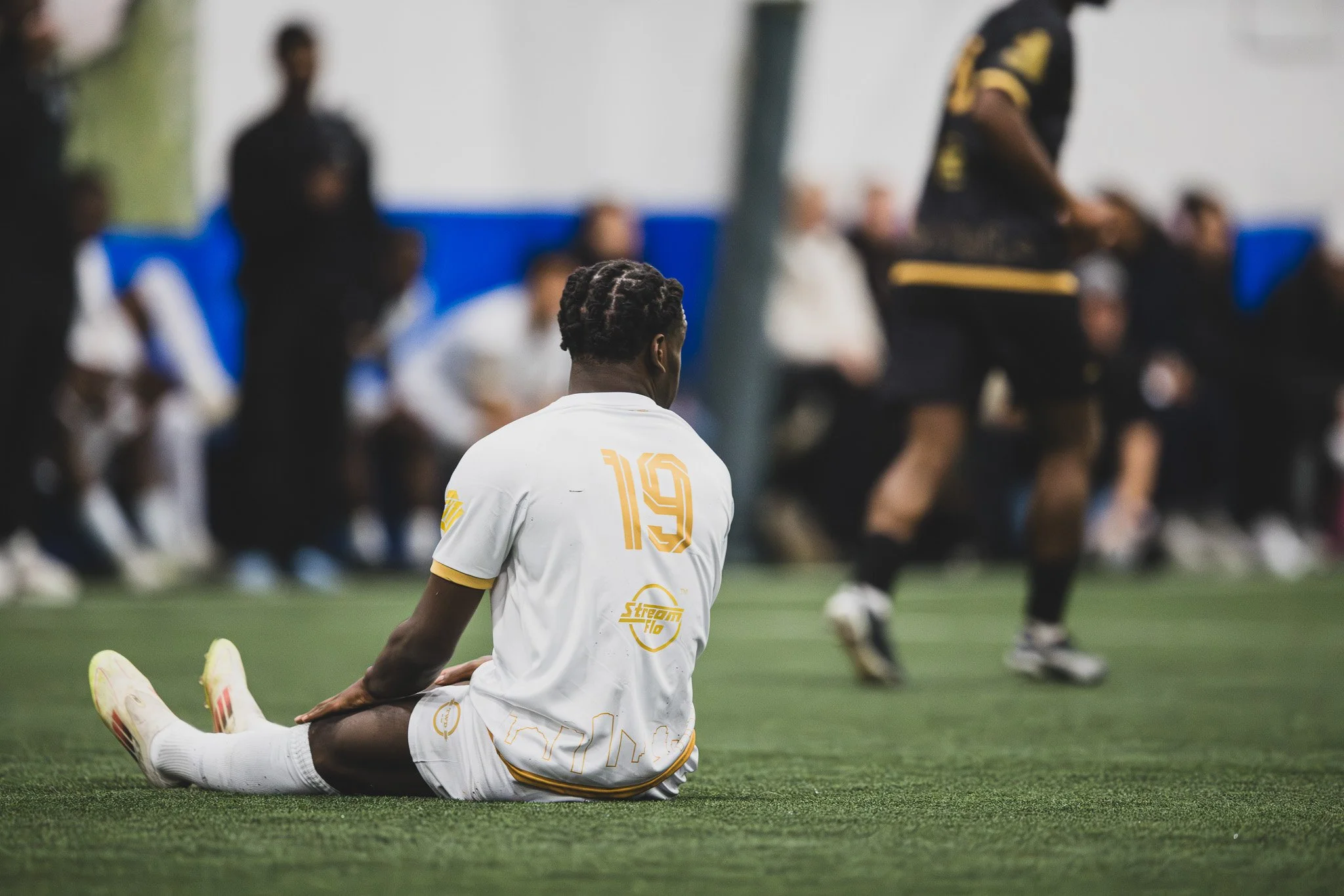 Athlete performing a split on the soccer field during a game or practice, with some spectators or team members seated in the background.