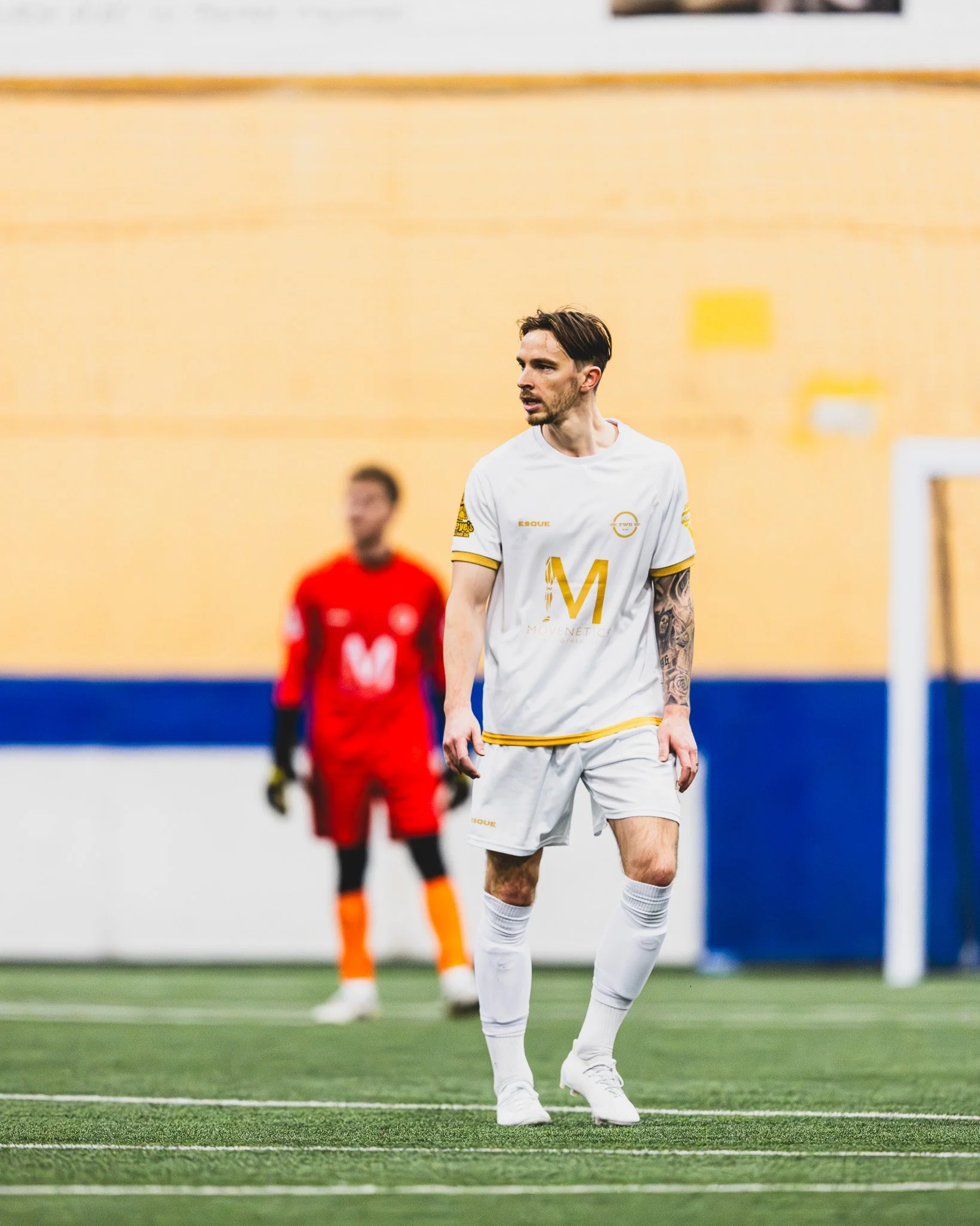 A soccer player wearing a white and yellow uniform walking on a green indoor soccer field with a yellow wall in the background. Another player in a red uniform is blurred in the background.