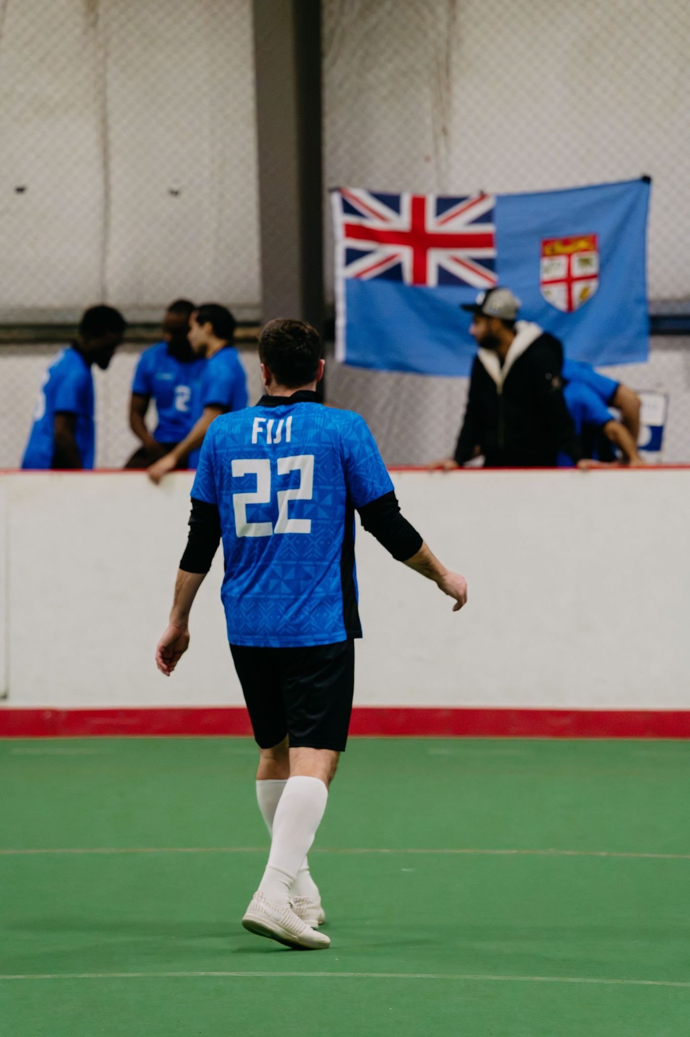 A soccer player wearing a blue jersey with the name 'FIJI' and the number 22 is walking on an indoor field. There is a group of people and a blue flag with a Union Jack and a crest hanging on the wall in the background.