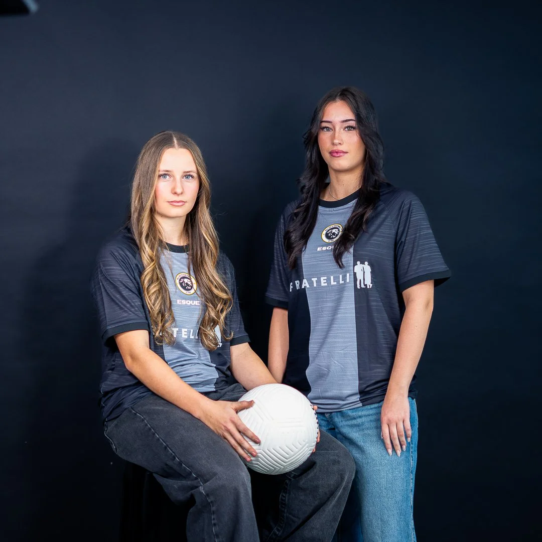 Two young women in esports jerseys, one sitting and holding a white volleyball, the other standing next to her, against a dark background.