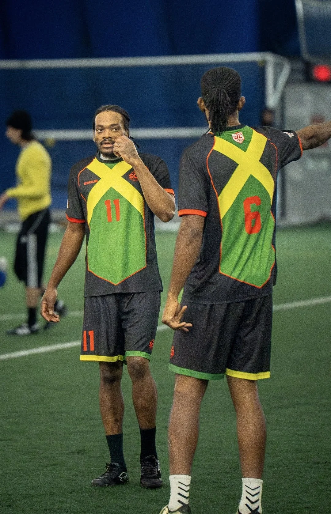 Two soccer players wearing matching black, green, and yellow jerseys with red numbers, standing on a field, preparing for a game.