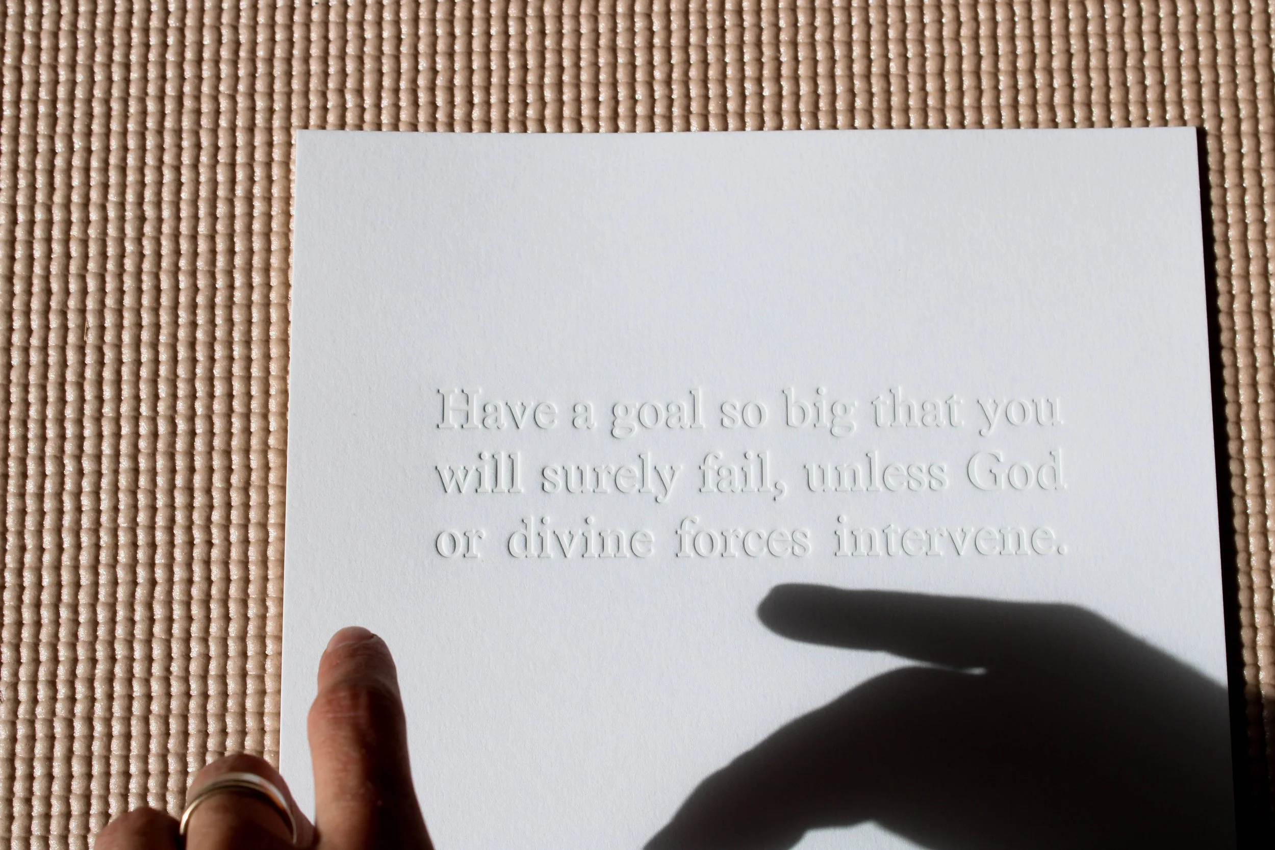 A white card with embossed text that reads, "Have a goal so big that you will surely fail, unless God or divine forces intervene." A person's hand with a gold ring on the finger is pointing at the card, casting a shadow on the text. The card is place