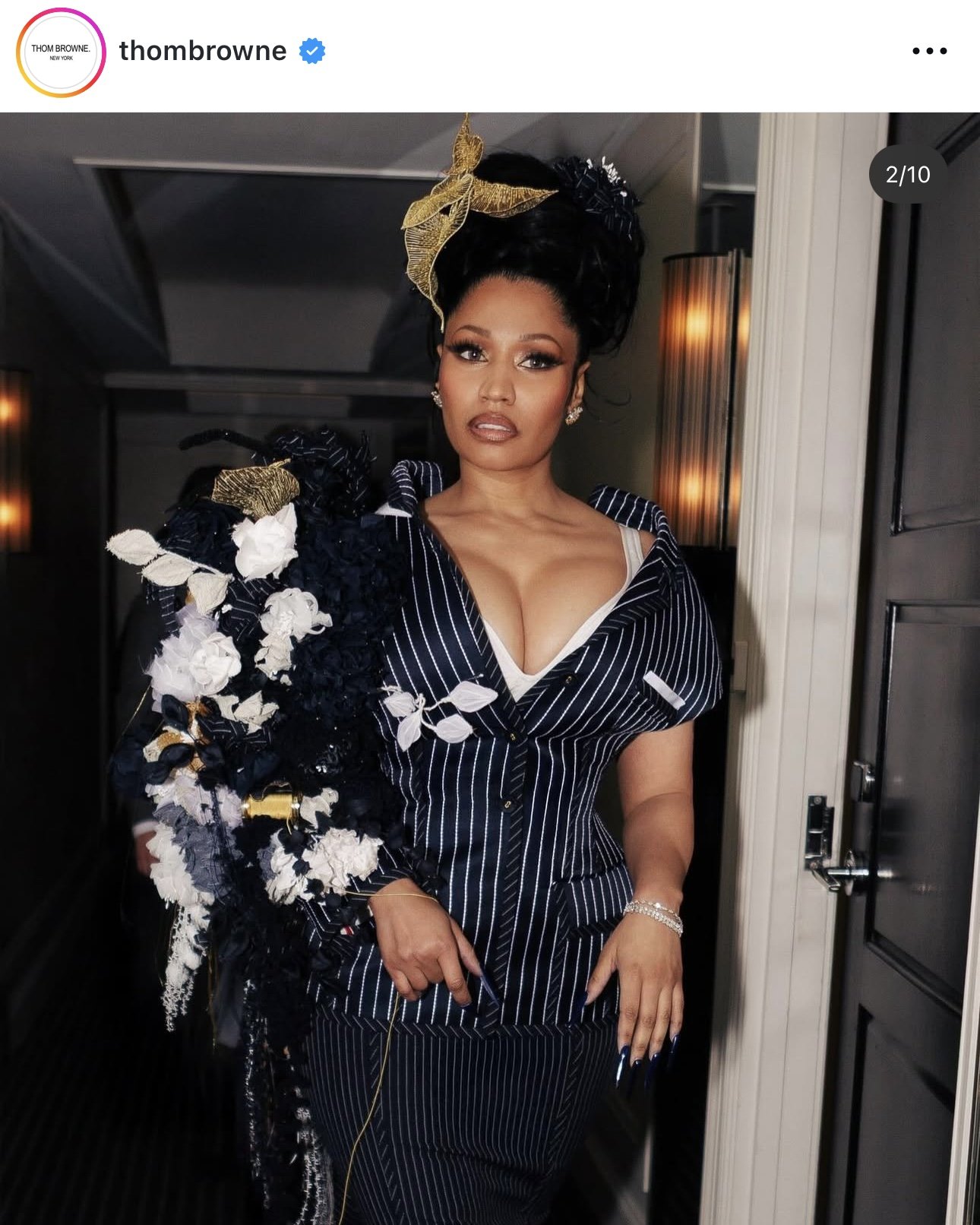 A woman dressed in a pinstripe suit with a floral embellishment on her shoulder, wearing earrings, a bracelet, and a gold leaf headpiece, standing in a dimly lit modern interior.