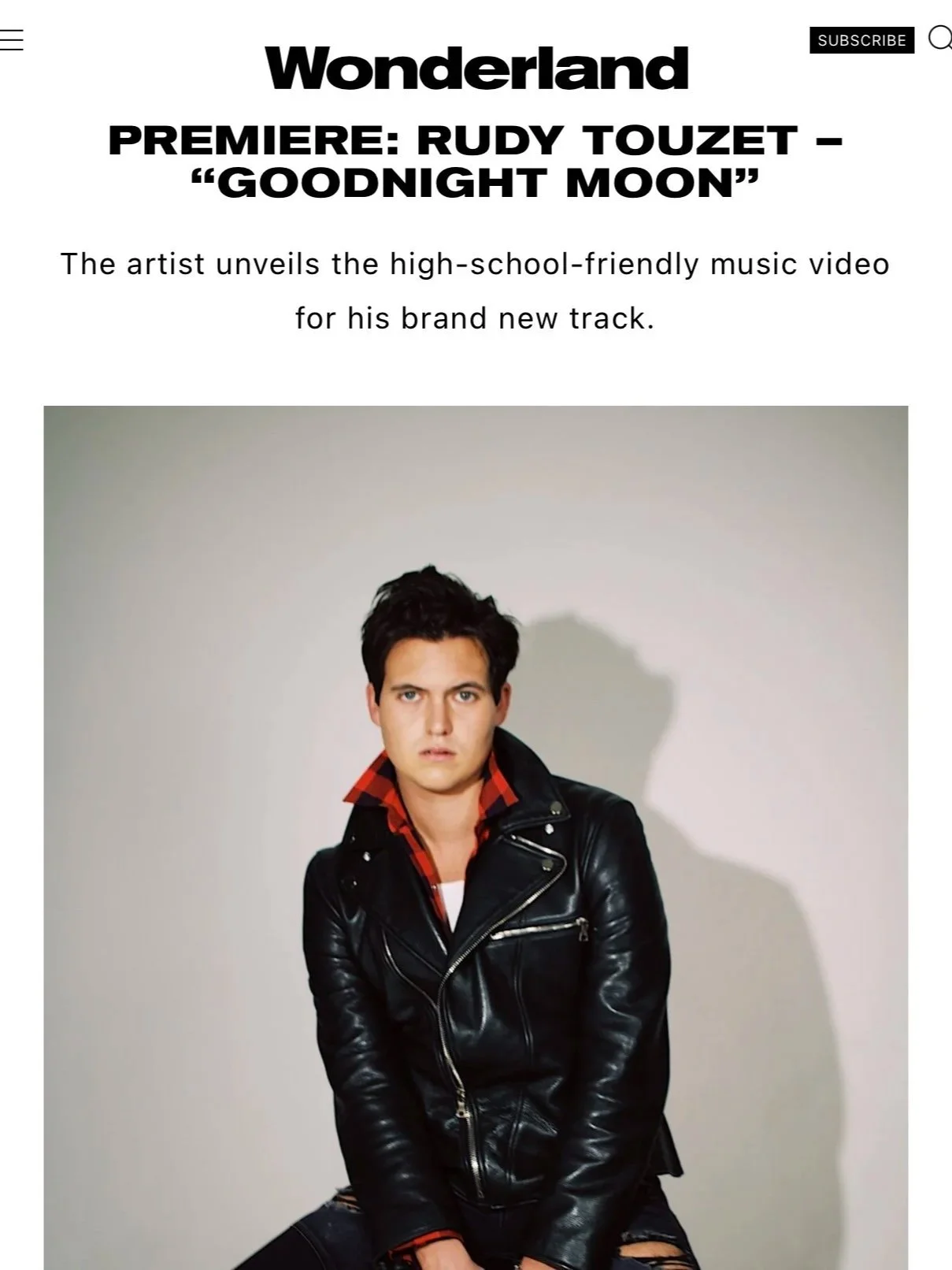 A young man in a black leather jacket with a red and black checkered shirt underneath, sitting against a plain white wall with a shadow cast behind him.