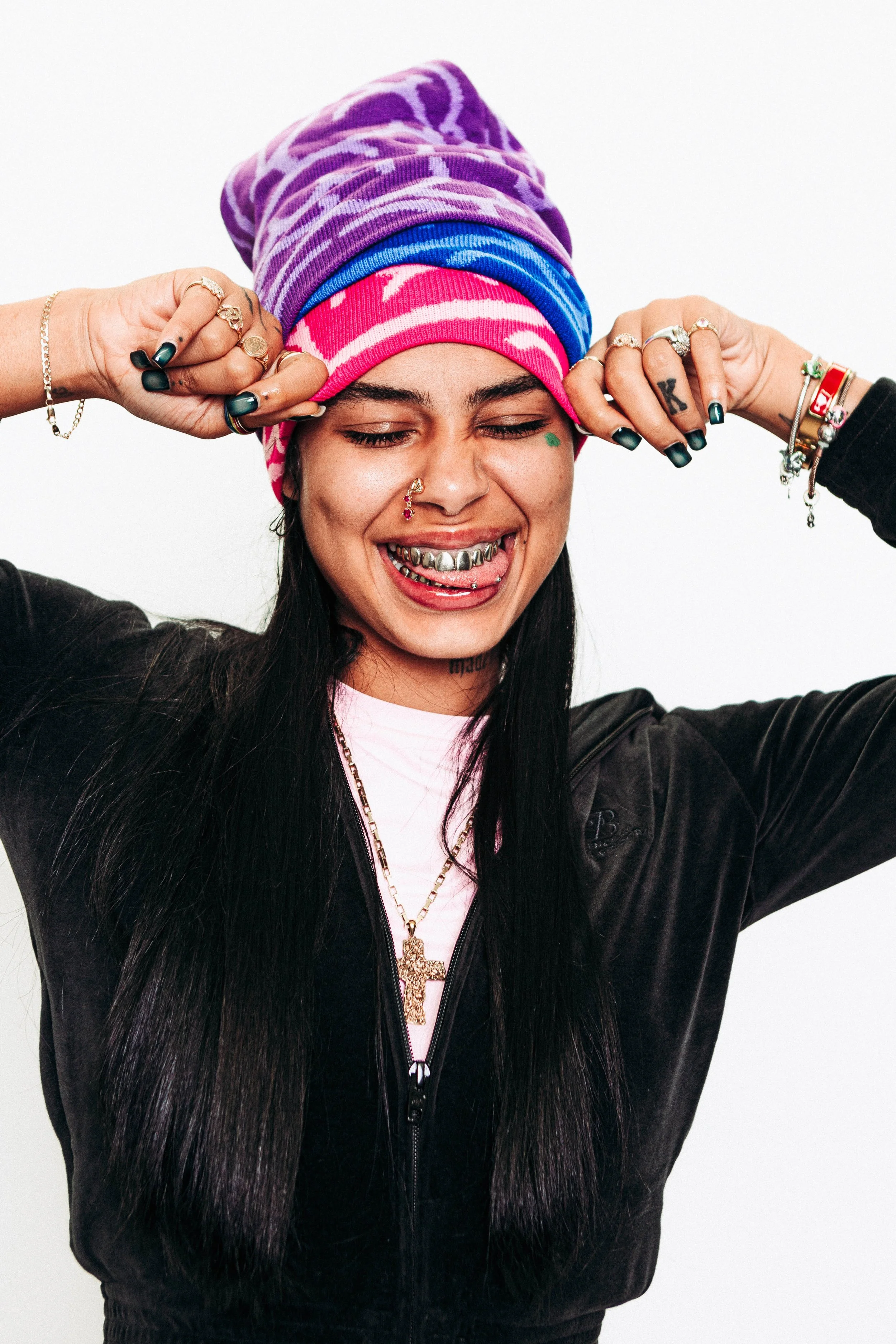 A young woman with long black hair wearing a colorful headscarf, multiple rings, bracelets, and a gold necklace, making a playful expression with her eyes closed and tongue out.