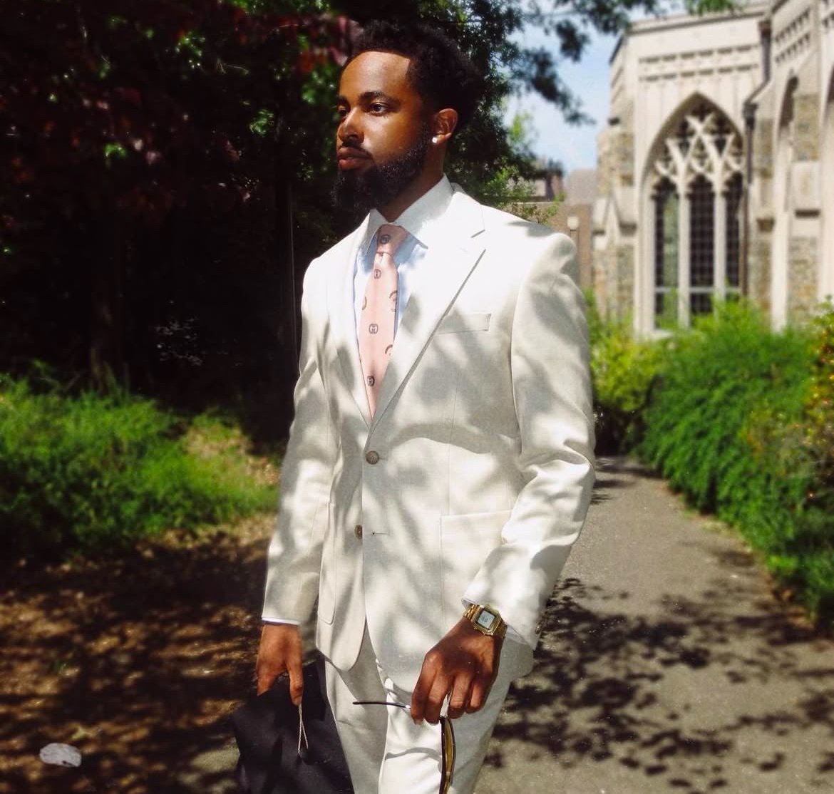 A man dressed in a white suit, pink patterned tie, and gold watch standing outdoors near a grassy area and historic building.