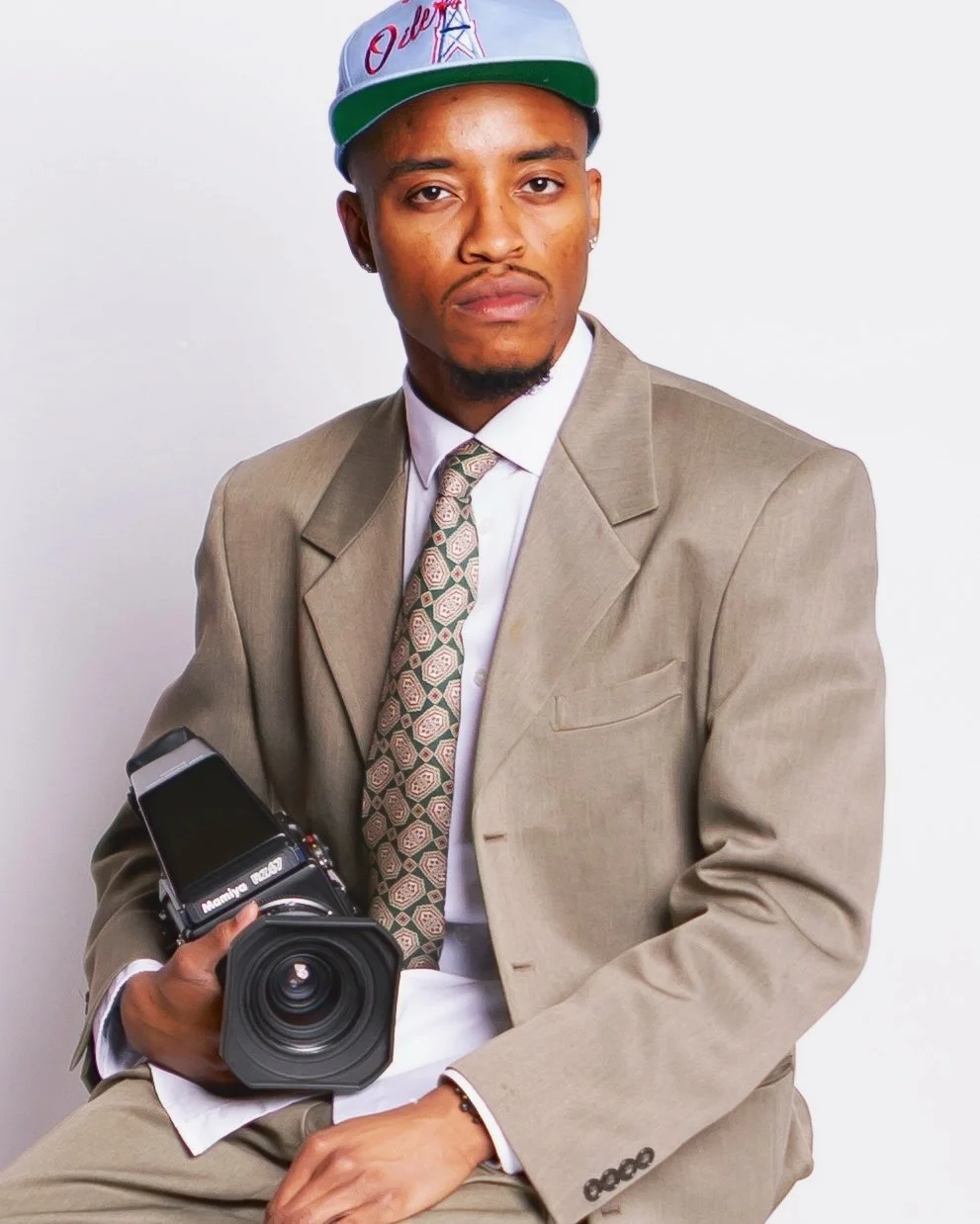 A young man in a beige suit, white shirt, and patterned tie, wearing a gray baseball cap with red and green accents, holding a professional camera, sitting against a plain white background.
