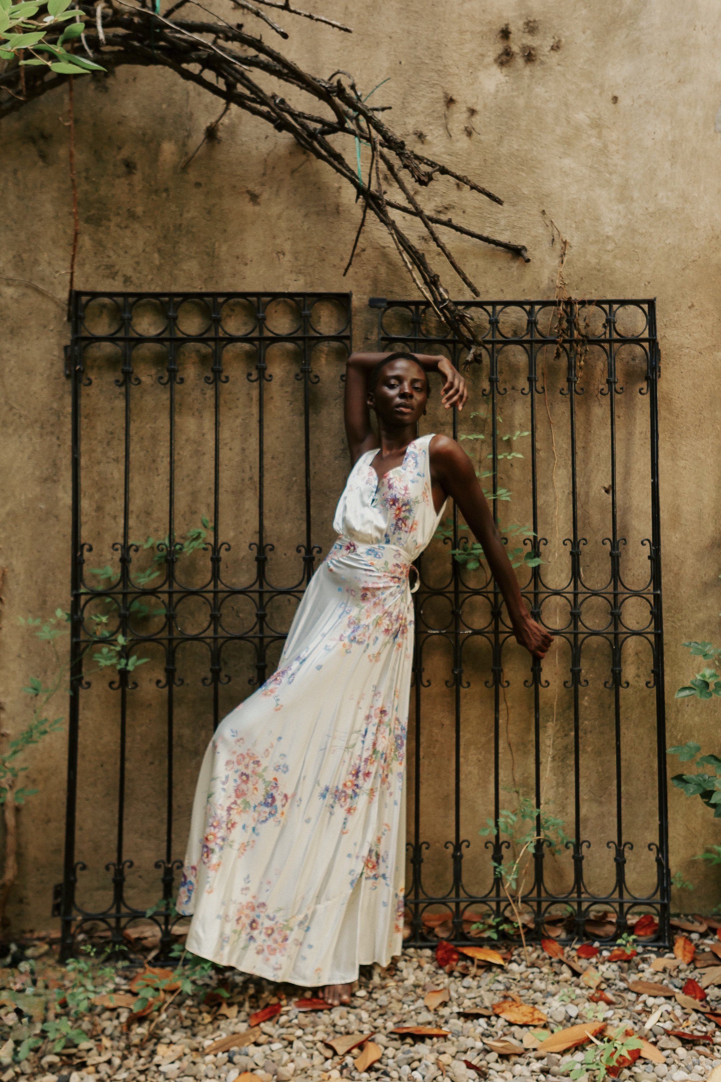 A woman in a long floral dress poses against a rustic beige wall with a black iron gate, surrounded by greenery and fallen leaves.