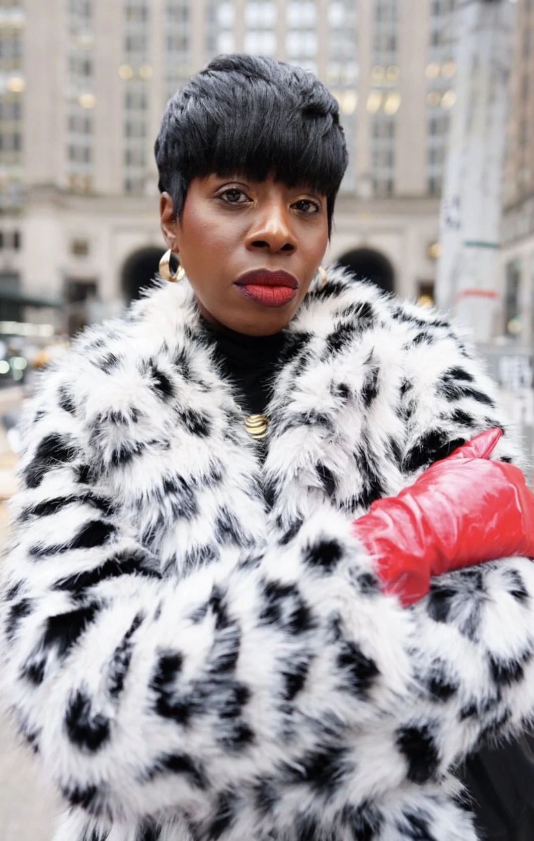 A woman with short black hair, wearing a faux fur coat with black and white spots, red gloves, gold earrings, a black turtleneck, and red lipstick, standing outdoors in an urban setting.