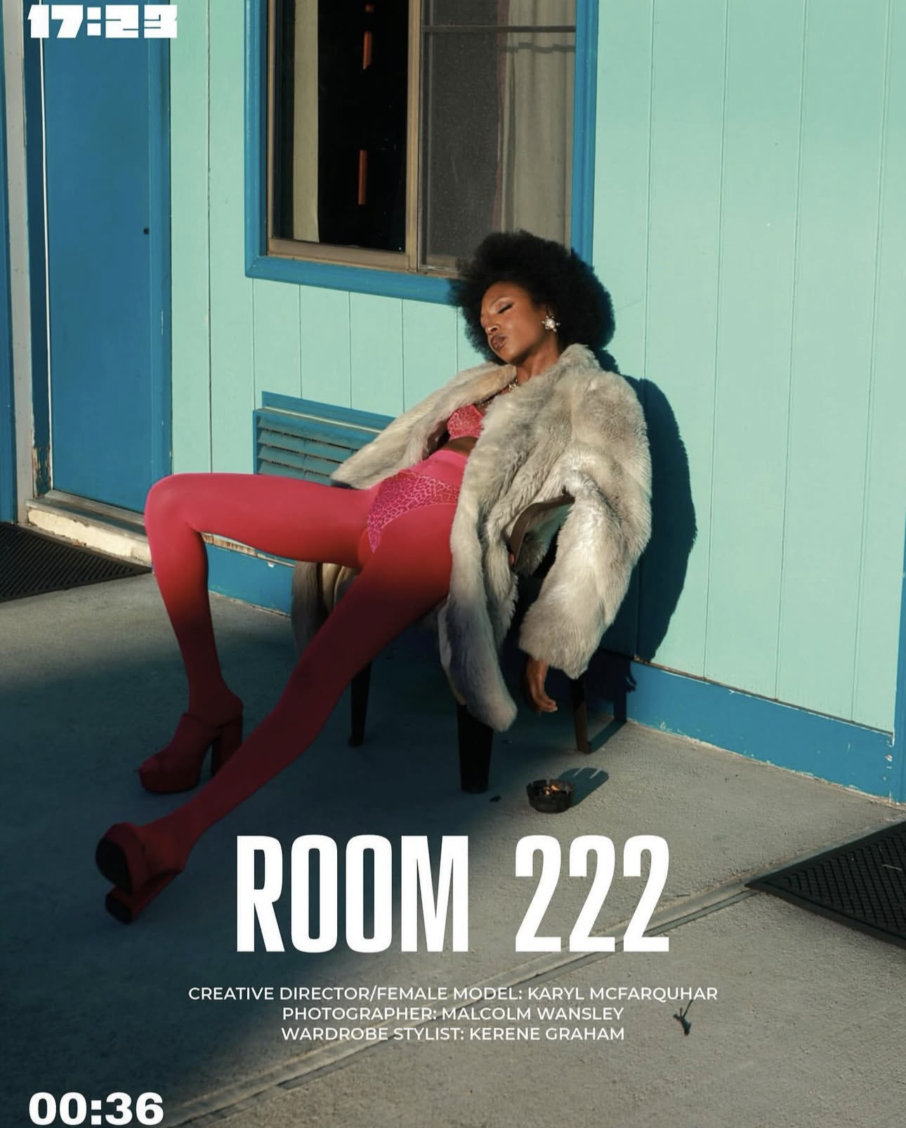 A woman with an afro hairstyle and dress in bright pink, wearing a fur coat, sitting on a chair outside a teal-colored building with a blue door and window, with her eyes closed and head tilted back.