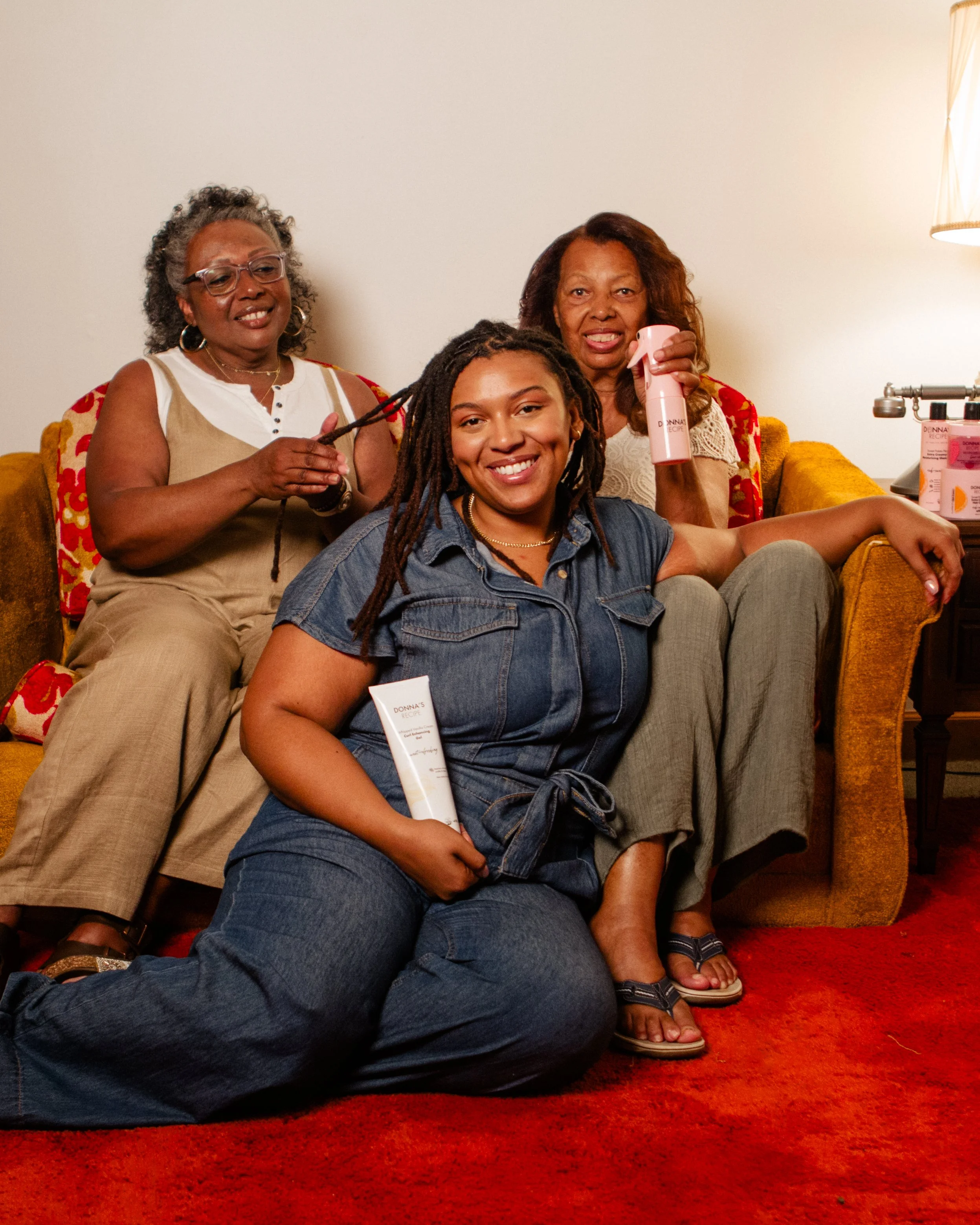 Three women with dyed hair and dreadlocks sitting on a sofa, smiling at the camera. One woman is in front, holding a tube of skincare product. The other two women are behind, one with a spray bottle and the other with a curling iron, in a cozy, warml