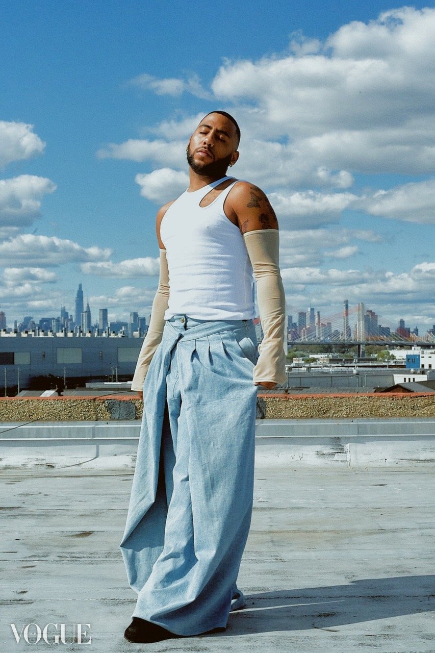 A man standing on a rooftop with New York City skyline in the background, wearing wide-leg jeans, a sleeveless white tank top, and beige arm sleeves, with arms relaxed and eyes closed.