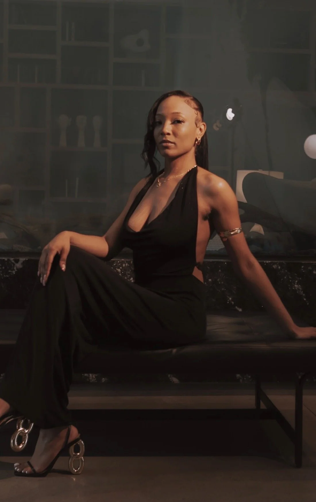 A woman in a black dress with side cutouts sitting on a black bench, wearing high heels, in a dimly lit room with a bookshelf and studio lights in the background.