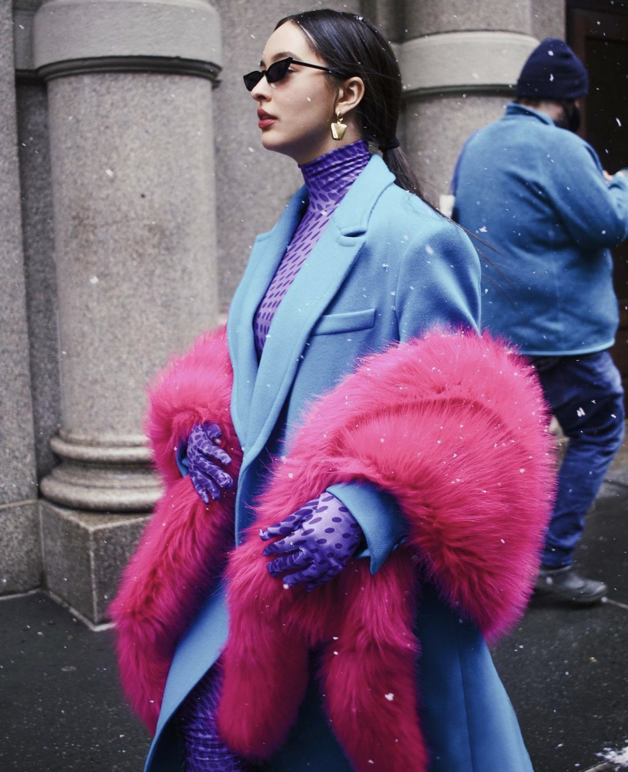 Woman wearing a blue coat with pink fur trim, purple patterned gloves, purple turtleneck, black sunglasses, and gold earrings, standing outdoors in winter with snow falling.