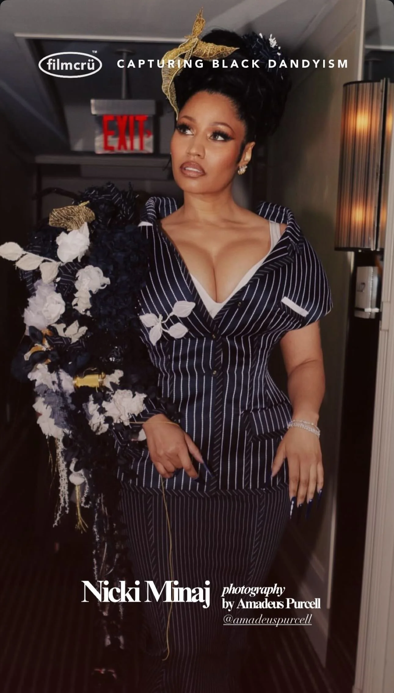 Nicki Minaj posing with a floral arrangement, wearing a pinstripe suit with a deep neckline, jewelry, and a headpiece, inside a room with an exit sign and wall lamp.