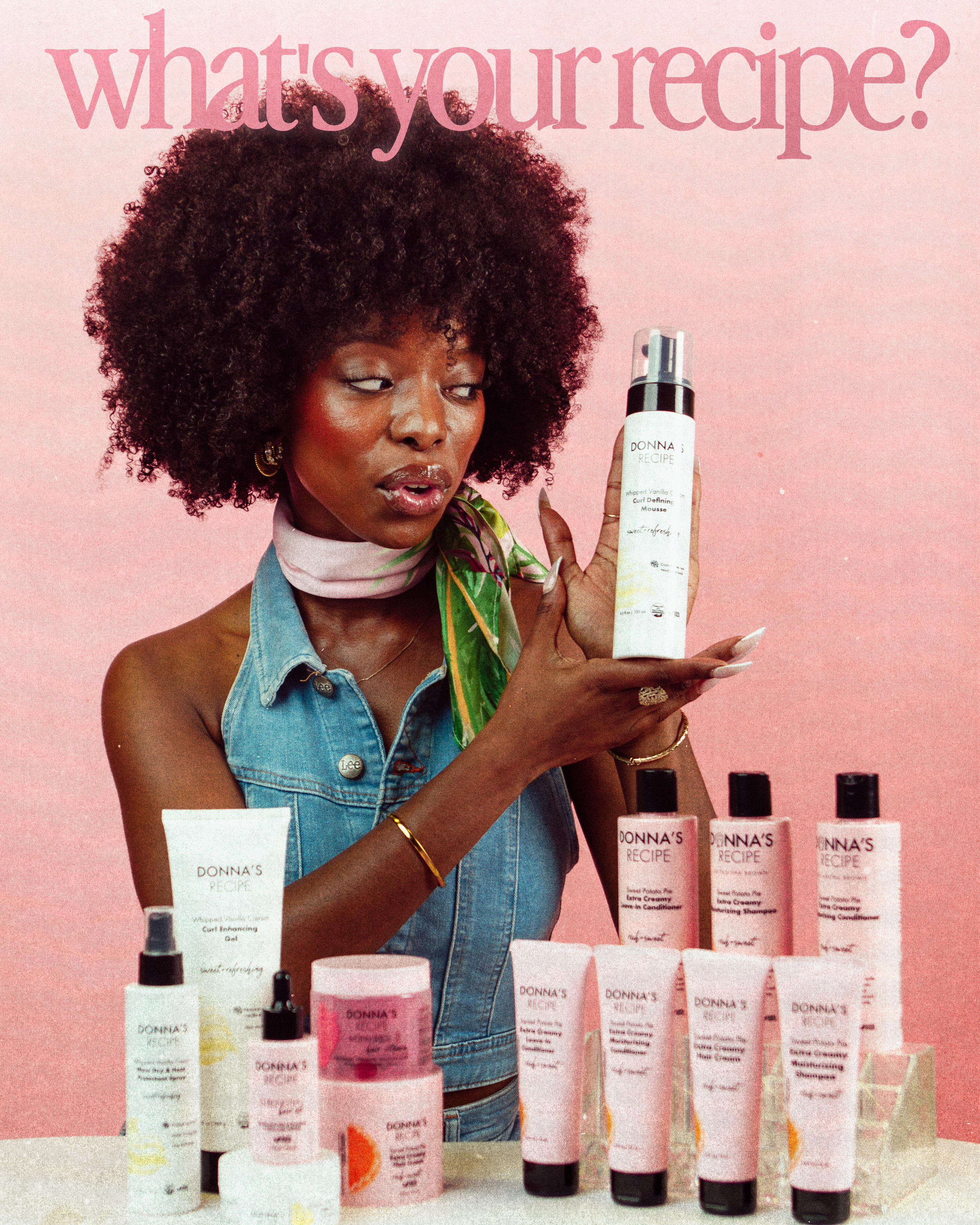 Woman with curly hair and denim vest holding a bottle from Donna's Recipe skincare line, surrounded by various skincare products on a table against a pink background.