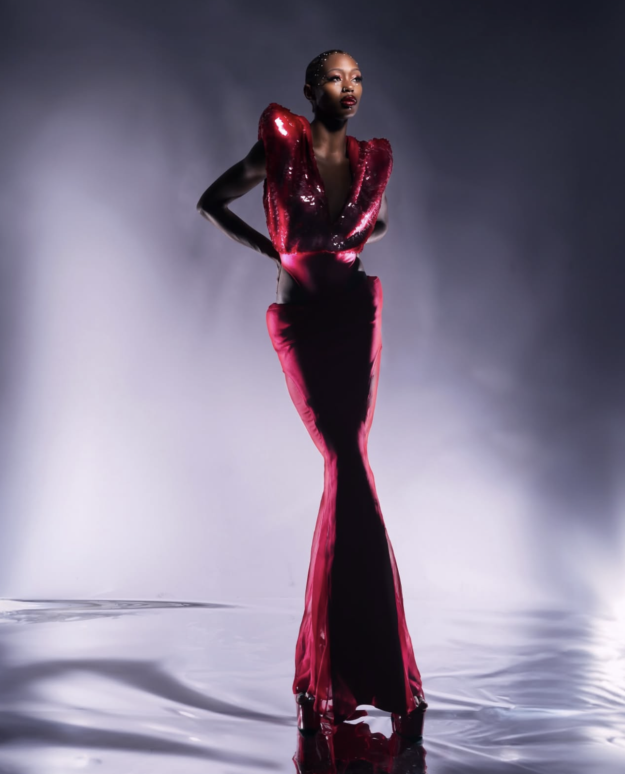 A woman in a glossy red gown with a deep neckline and padded shoulders poses against a dark background with soft lighting.