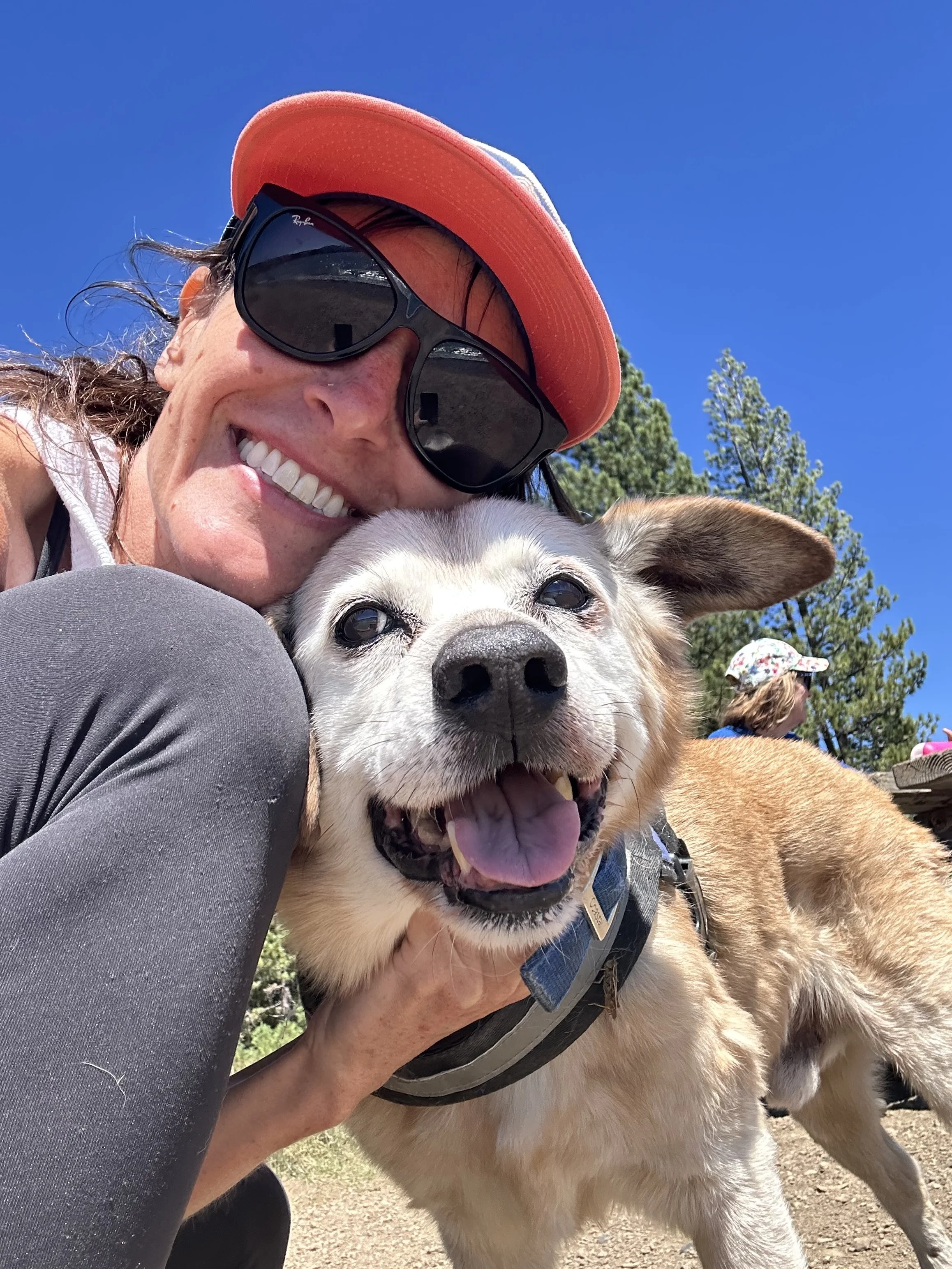 A woman smiling, wearing sunglasses and a red cap, hugging a happy dog outdoors on a sunny day with trees and a person in the background.
