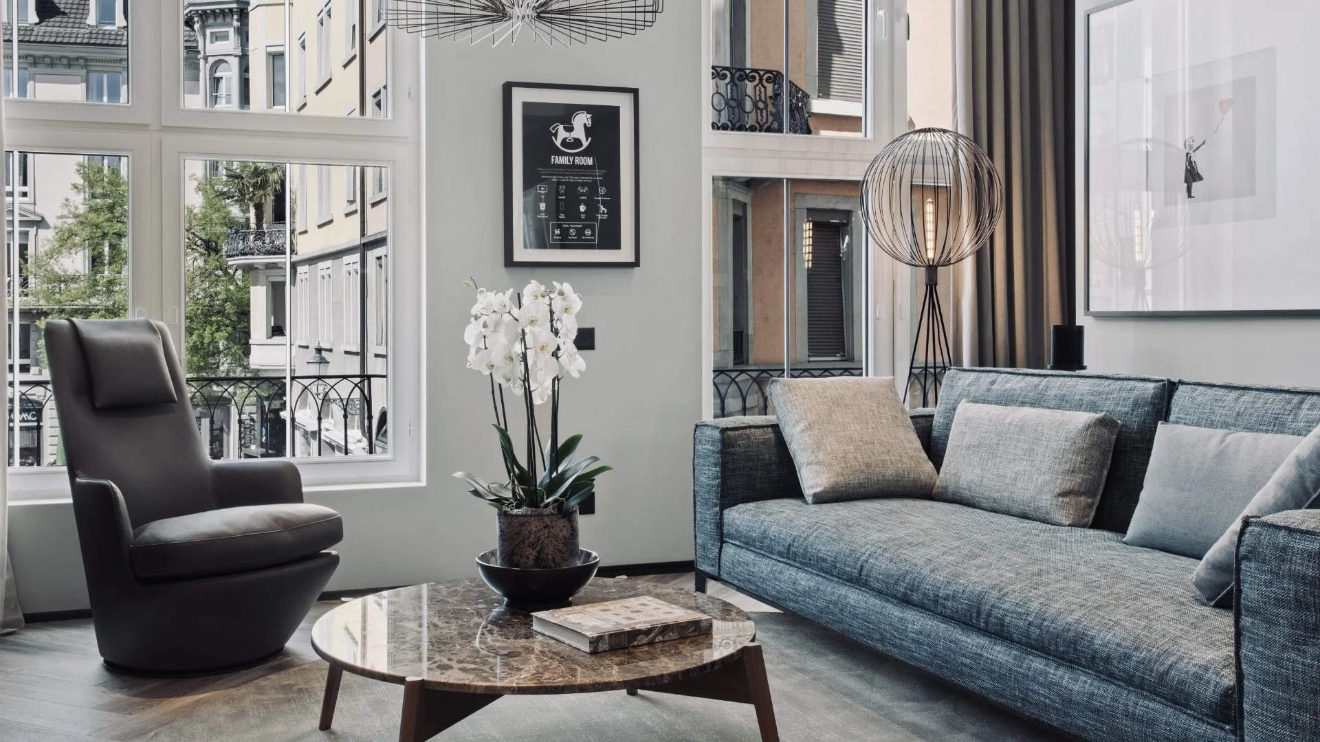 Modernes Wohnzimmer mit großen Fenstern, grauem Sofa, schwarzen Armstuhl, Couchtisch mit Buch und weißer Orchidee, Wandkunst und Stehlampe.