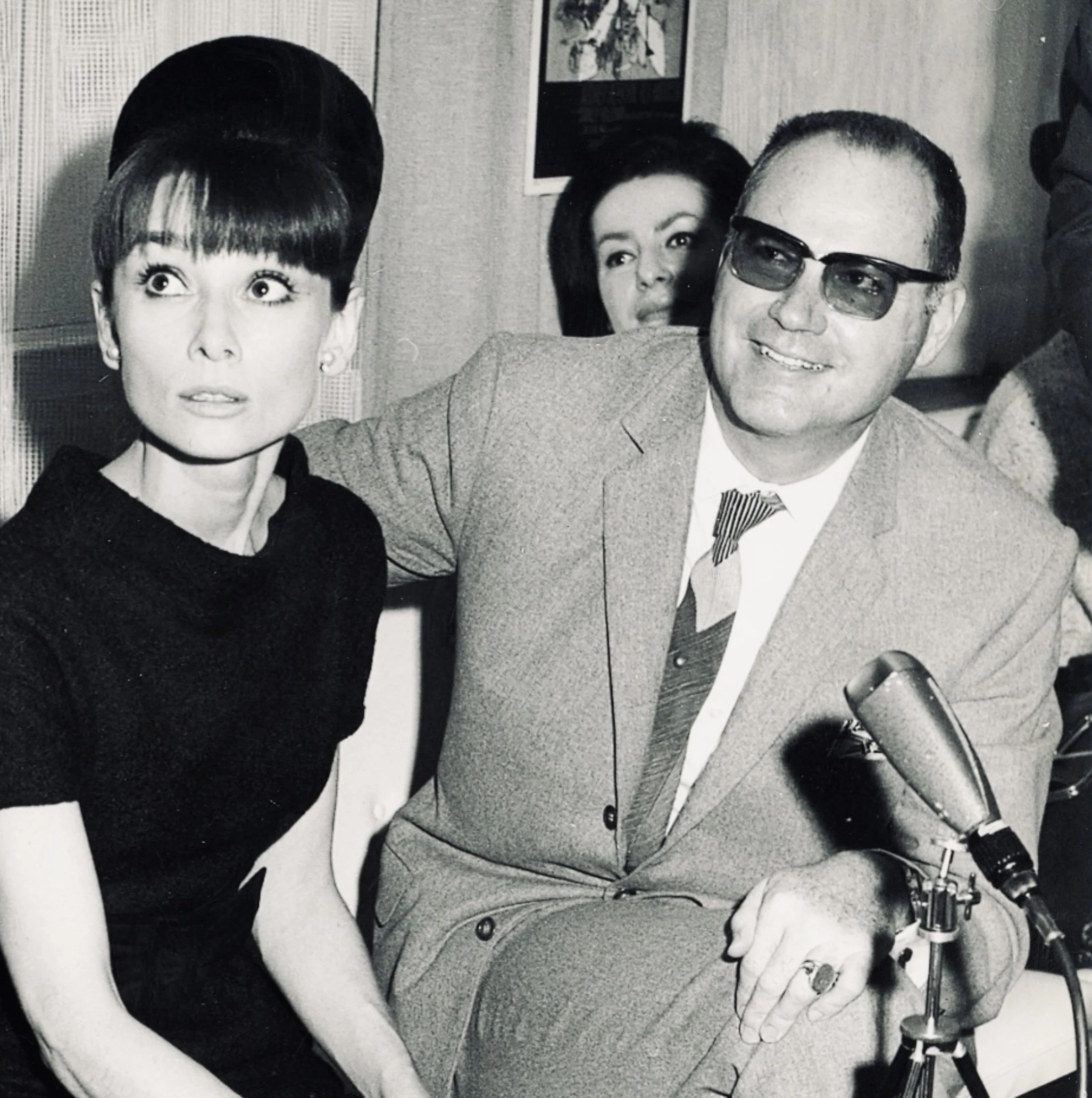 Schwarz-weiß-Foto von drei Menschen bei einer Radiosendung. Ein Mann trägt eine Brille und einen Anzug mit Krawatte, sitzt neben einem Mikrofon. Zwei Frauen sitzen hinter ihm, eine mit blonder Frisur und eine mit dunklen Haaren.