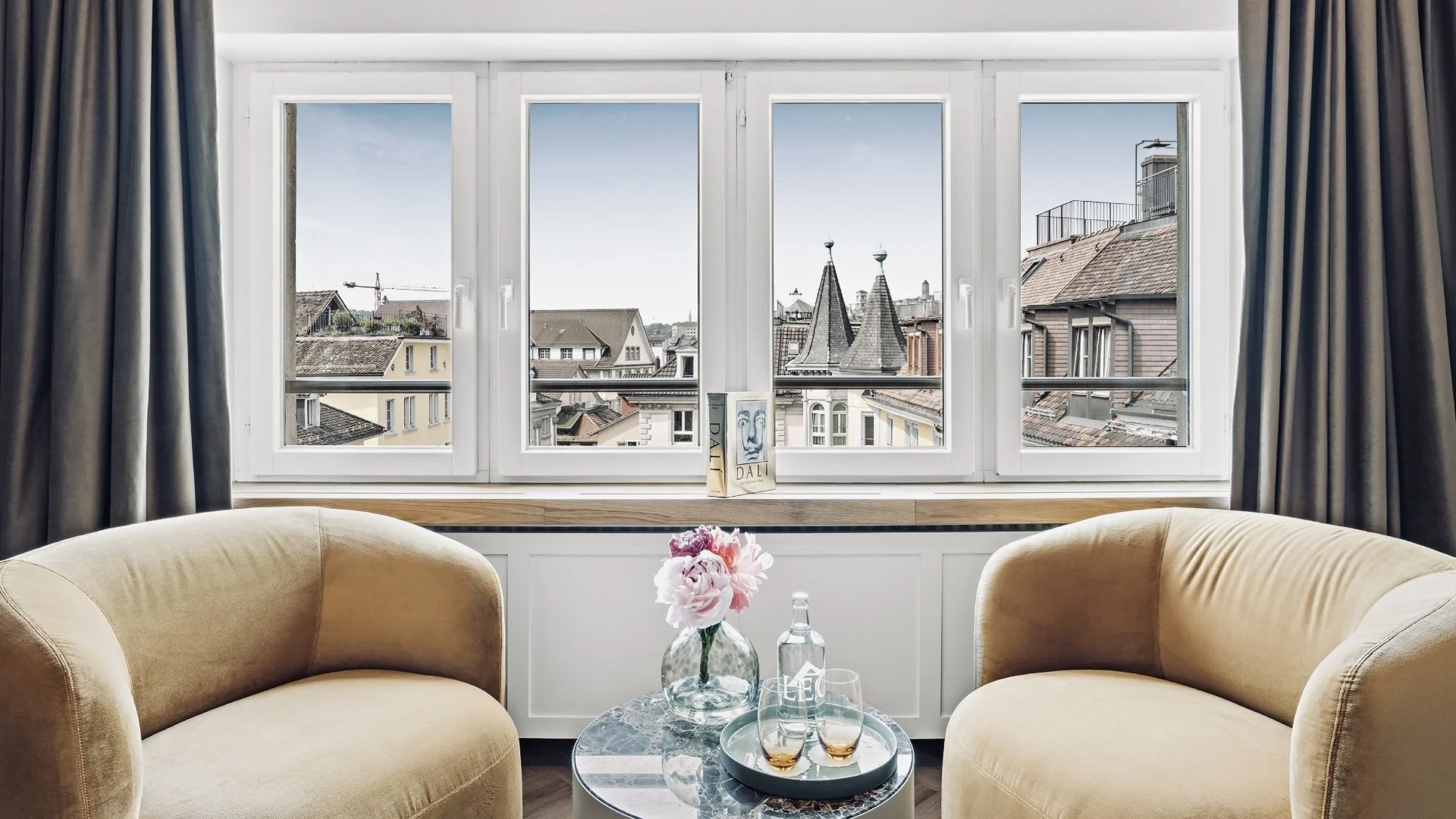 Gemütliches Wohnzimmer mit zwei beige Sofas, einer runden Marmor-Couchtisch mit einer Vase mit pinken Blumen, einer Flasche Wasser und zwei Gläser. Großes Fenster bietet Blick auf das Dach der Nachbarhäuser und Türme in der Ferne.