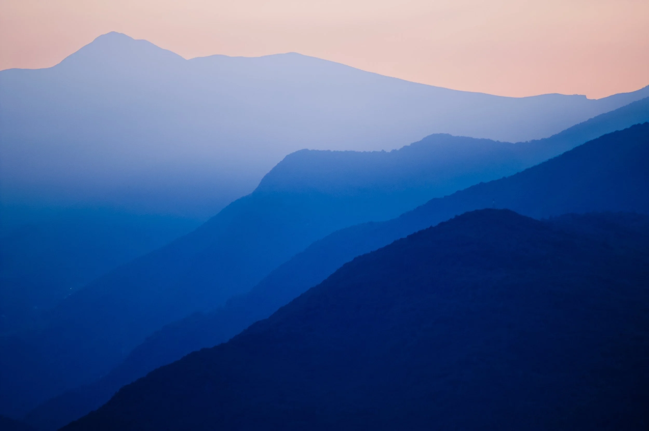 Layered mountain silhouettes fading from dark to light blue beneath a peach-toned sky, used to represent existential therapy.