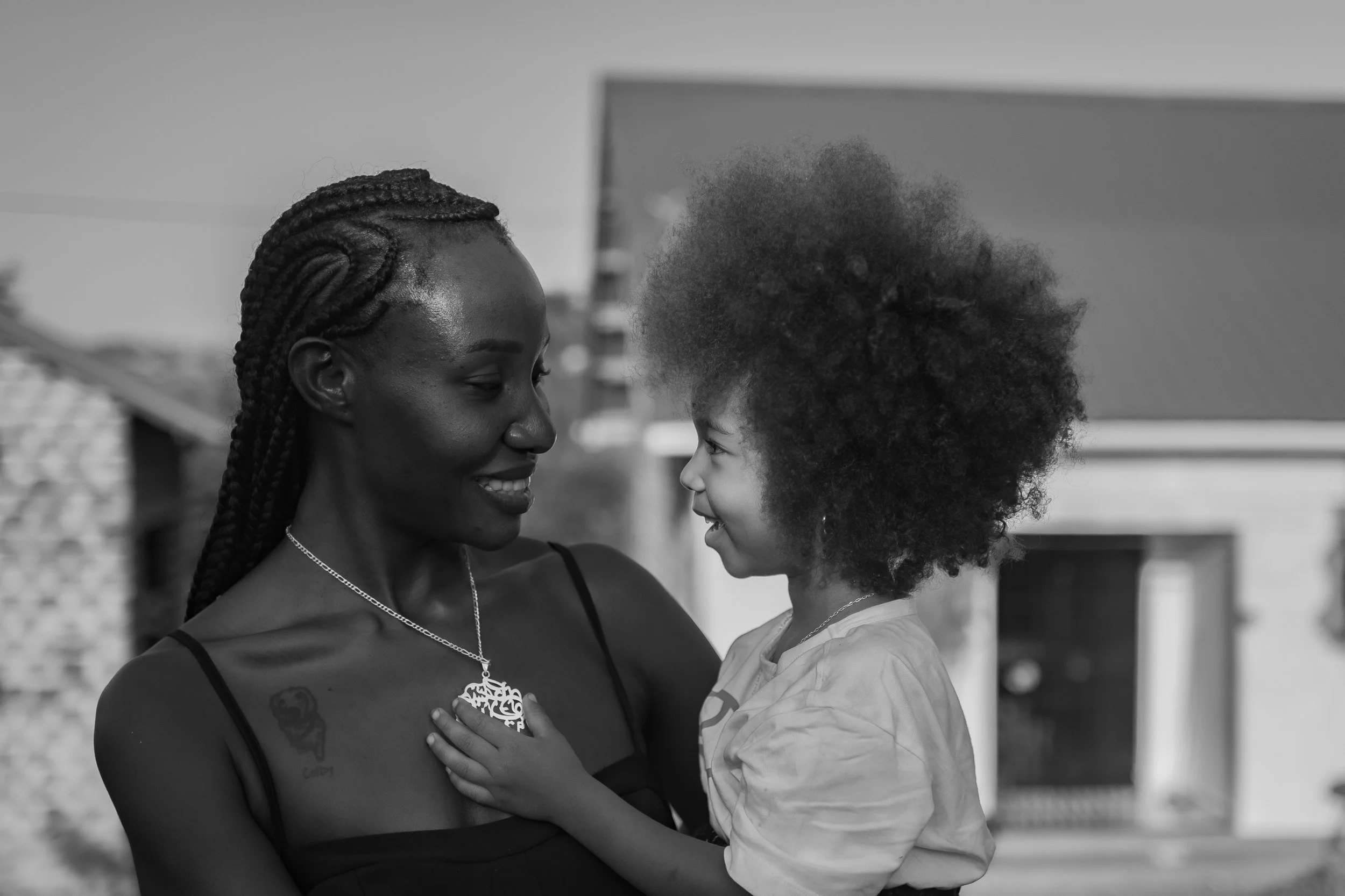 A woman with braided hair and a tattoo on her shoulder holding a young girl with curly hair and smiling at her outside