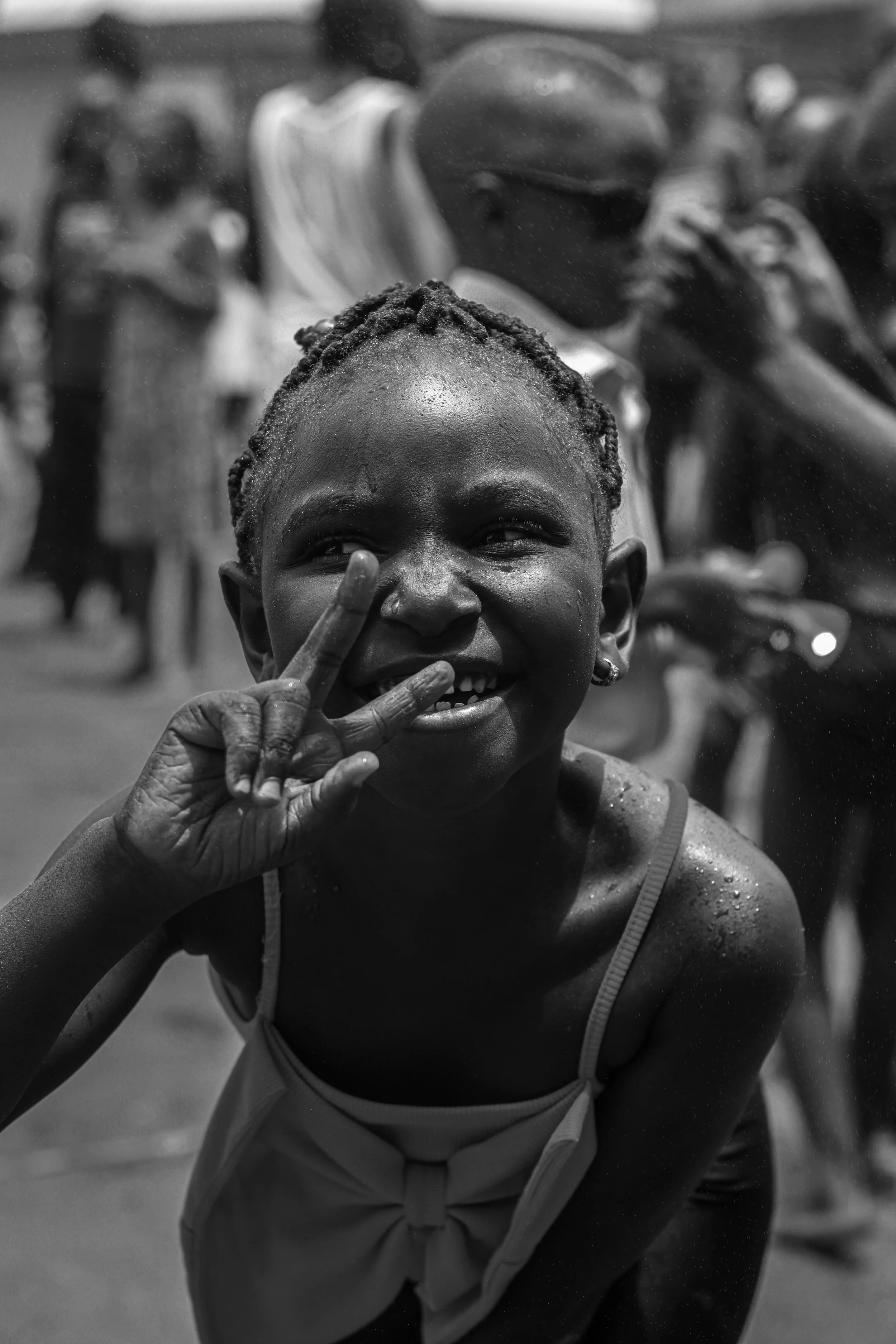 A young girl smiling and making a peace sign with her fingers, surrounded by a crowd in the background, water on her face, in black and white.