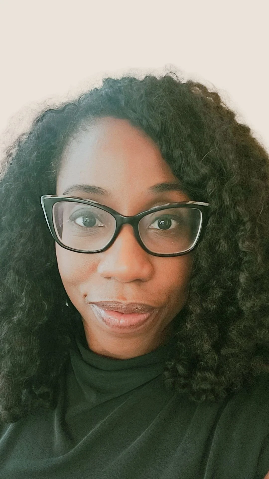 A woman with curly black hair, glasses, and a black top looking at the camera.