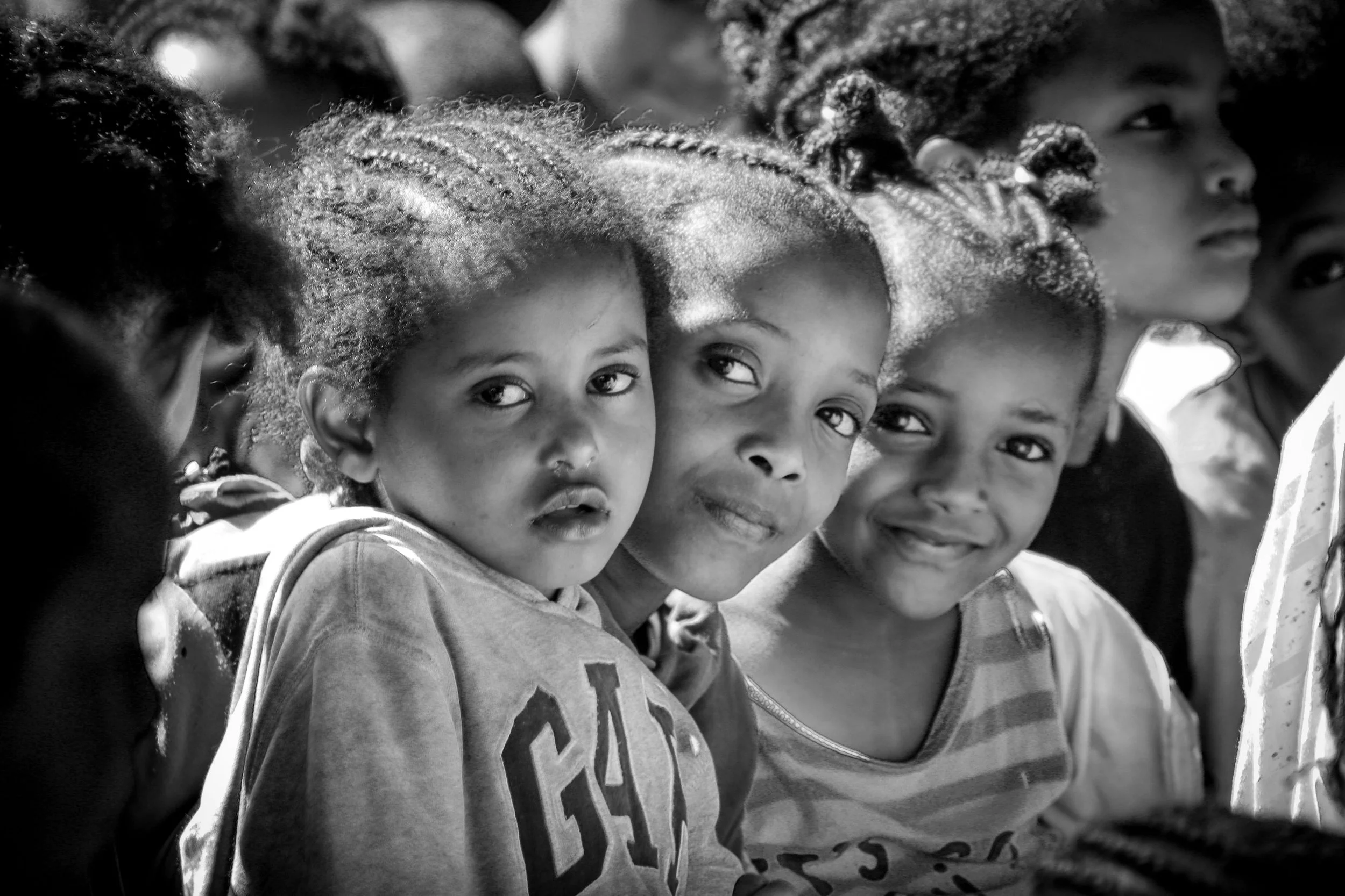 Group of young children, including girls with braided hair, sitting closely together and looking towards the camera, in black and white.