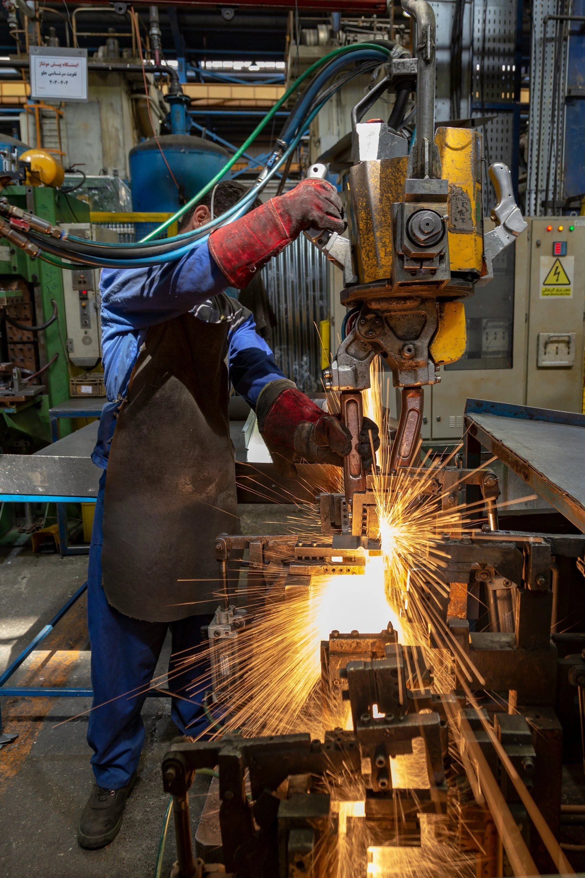 A worker in protective gloves and attire welding metal with sparks flying in an industrial workshop filled with machinery and equipment.