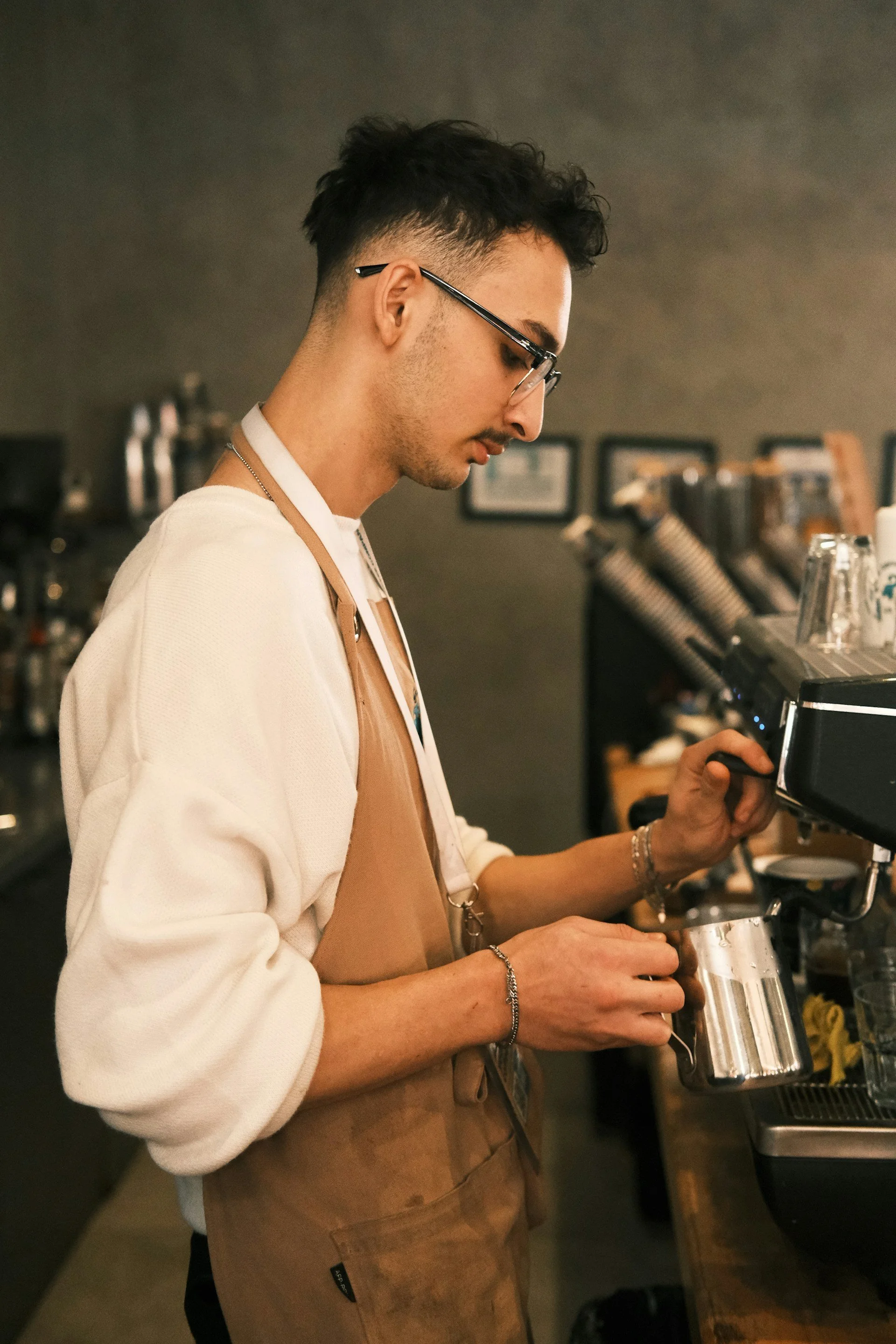 A barista wearing glasses and a brown apron making coffee in a coffee shop.