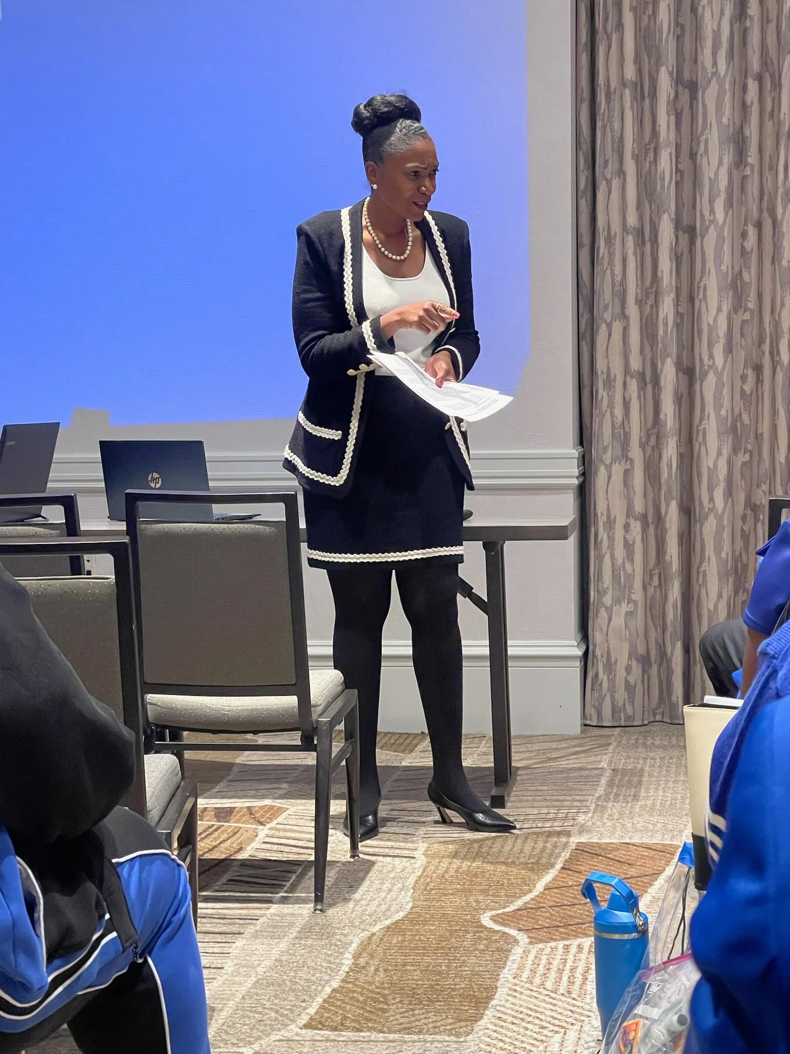 A woman with gray hair styled in a bun, wearing a black suit with white trim, a white blouse, pearl necklace, and earrings, standing and speaking to a group in a conference room. She is holding papers in her left hand and gesturing with her right hand.