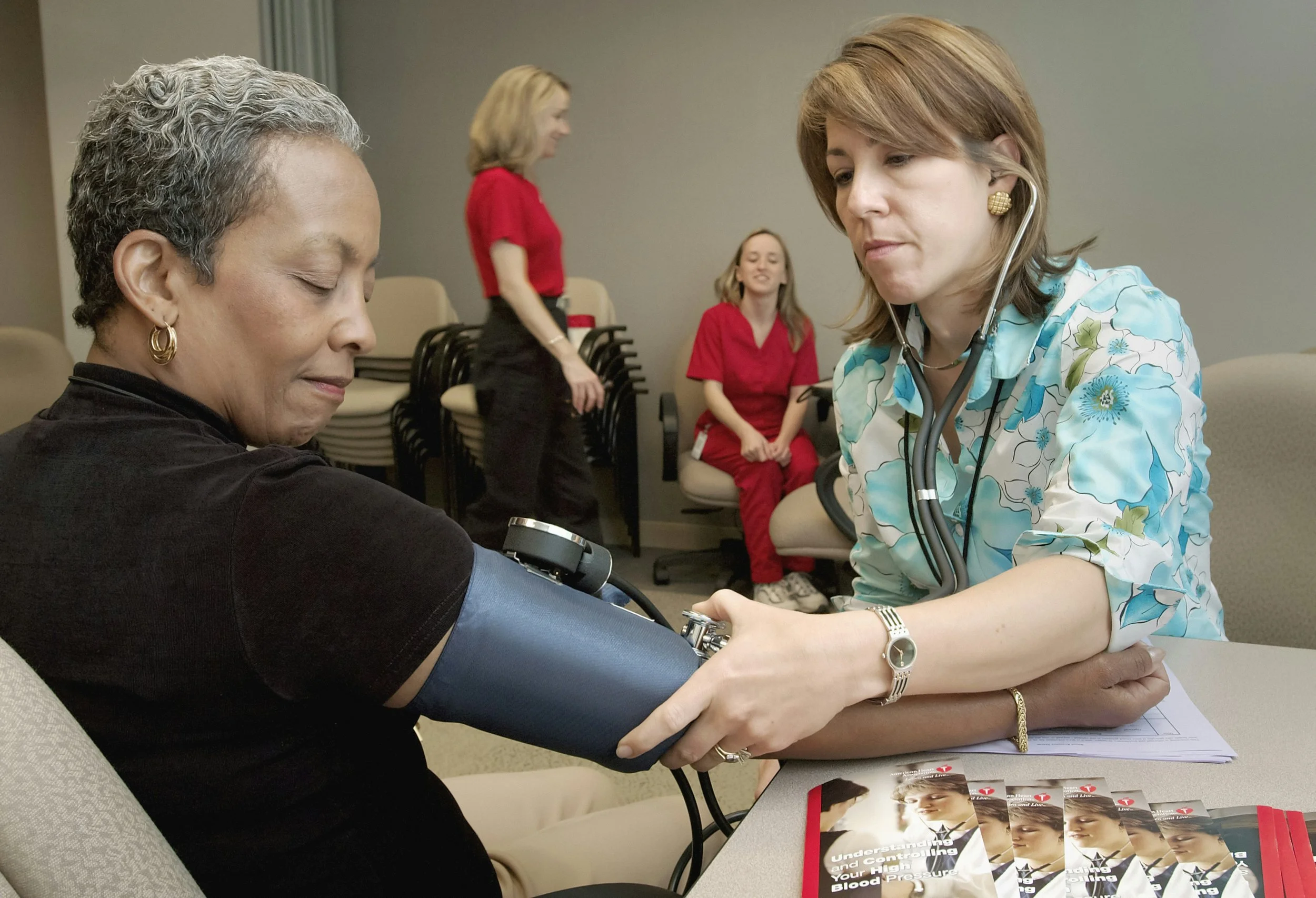 A healthcare professional measures a woman's blood pressure in a medical setting with two other women observing.