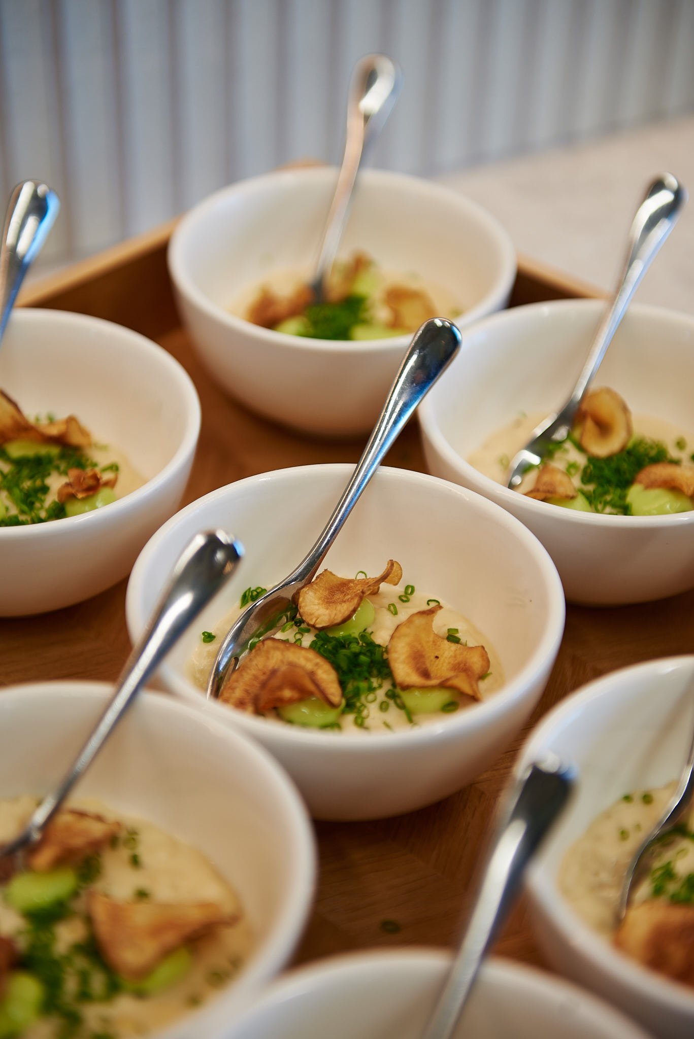 Several small white bowls filled with a creamy dish topped with green herbs and crispy garlic slices, each with a silver spoon inside, arranged on a wooden tray.