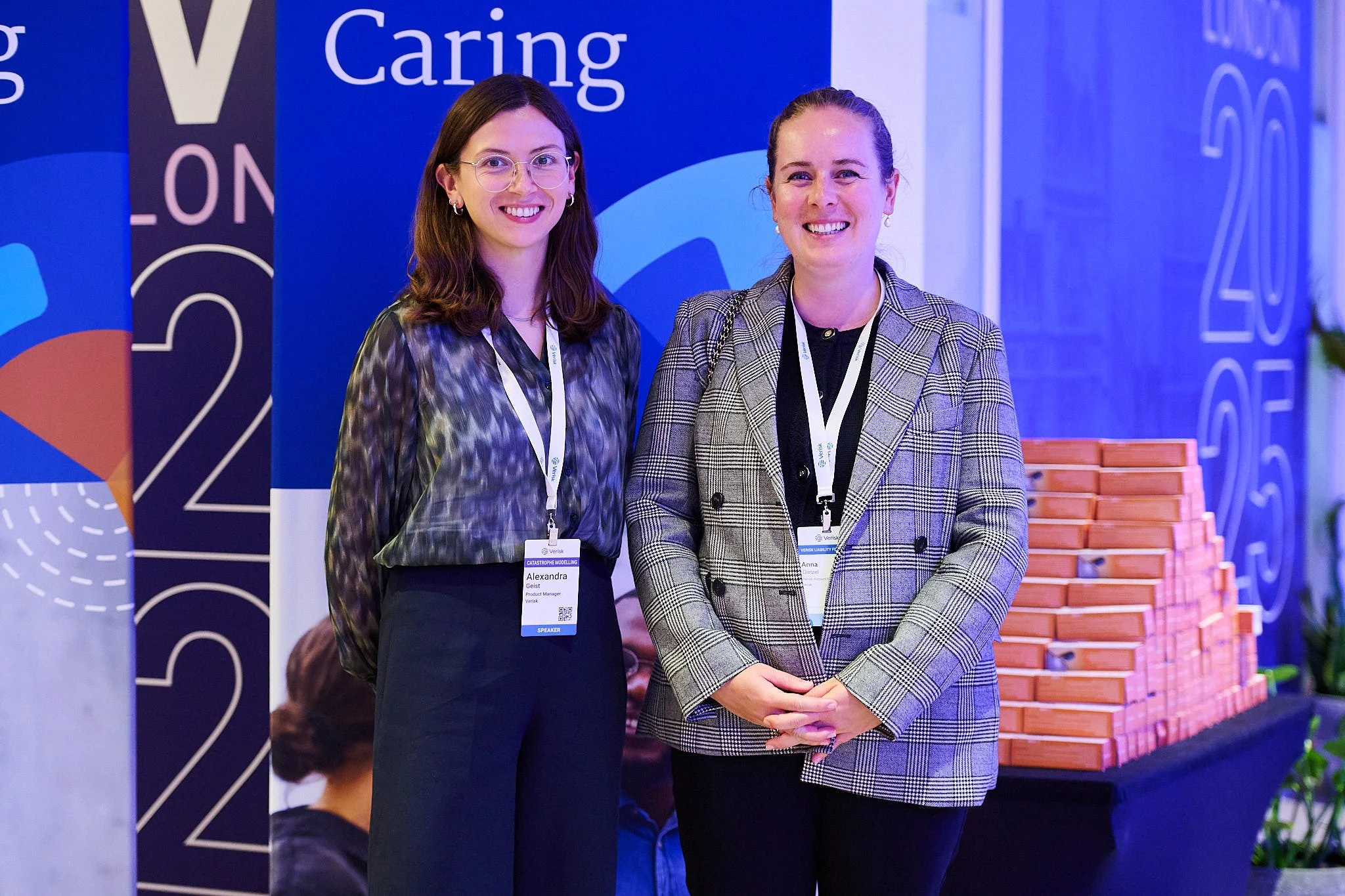 Two women smiling at a conference or event, wearing name tags, standing in front of a blue background with words and graphics