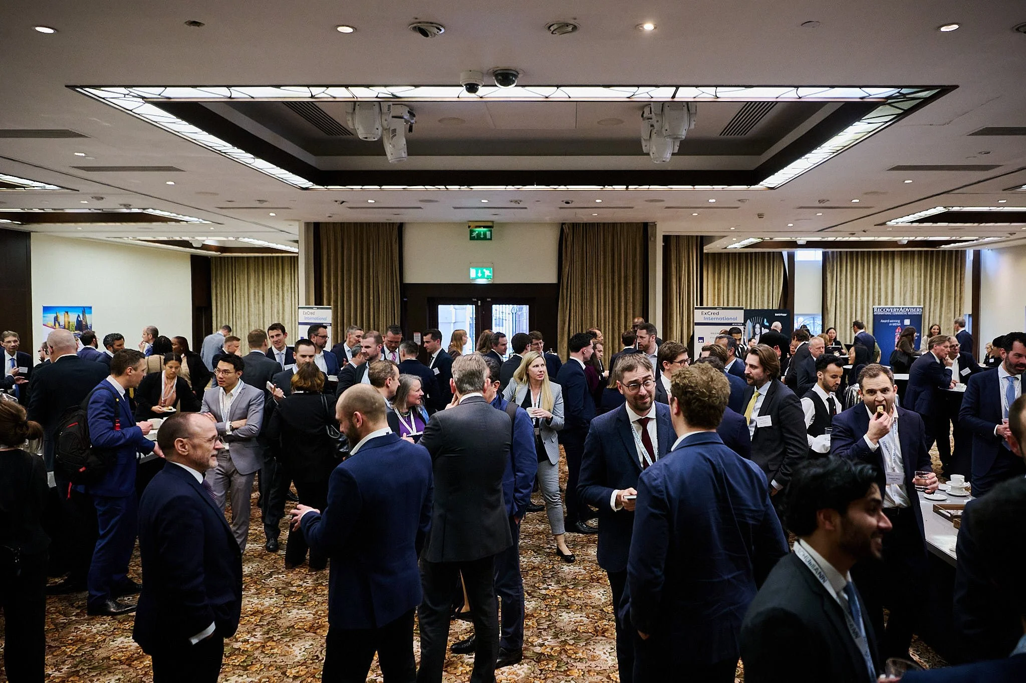 A busy networking event or conference with many professionals in formal business attire, chatting and mingling in a large, well-lit conference room.
