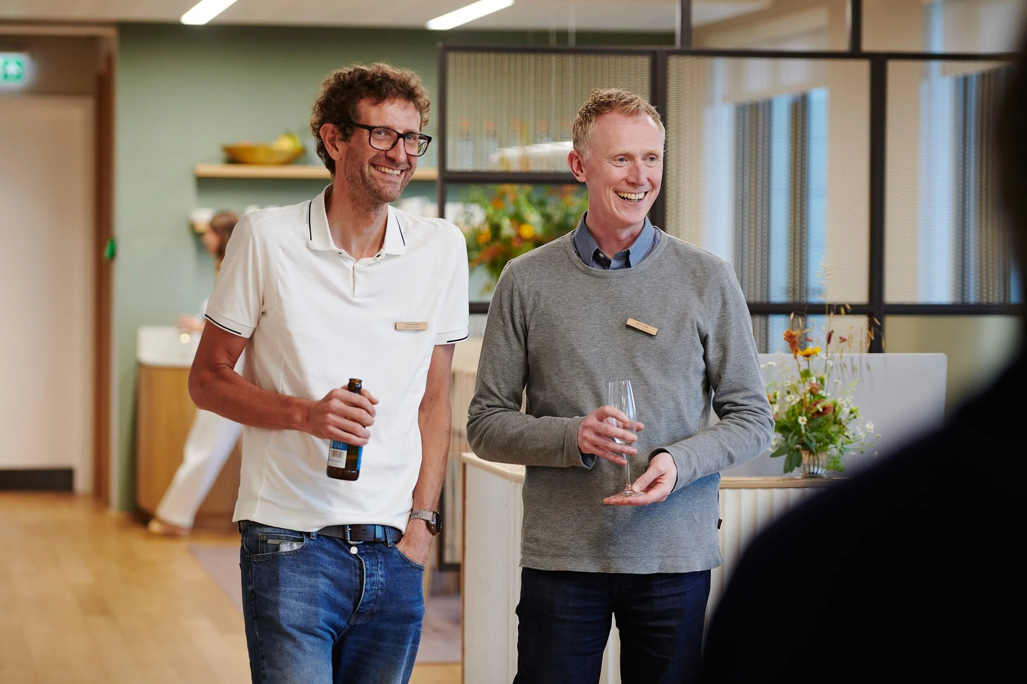 Two men smiling and talking at a social gathering, one holding a bottle and the other holding a wine glass, in a modern indoor setting with flowers in the background.