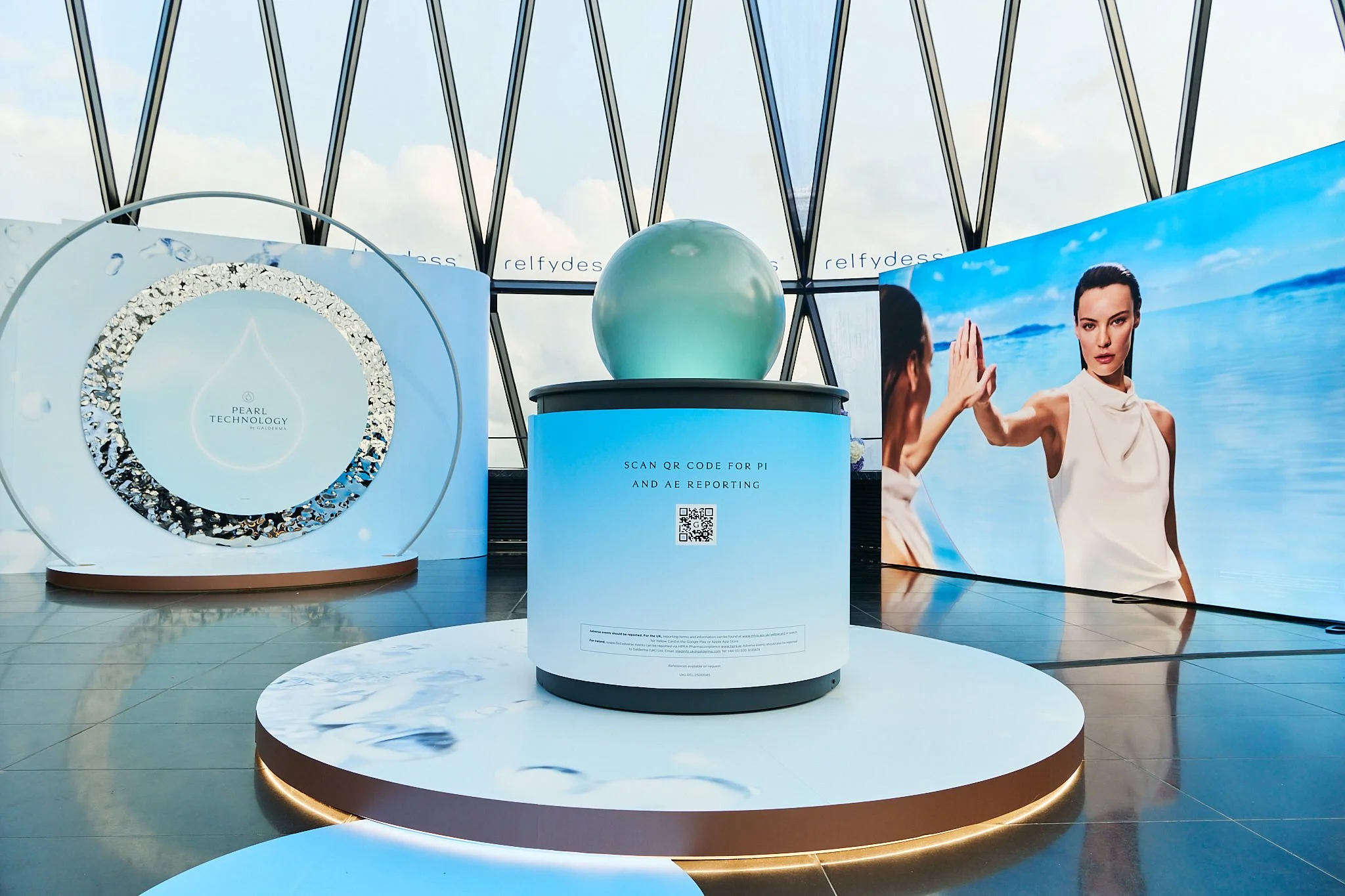 A modern exhibit display with a large pearl-shaped sculpture on a blue stand in a bright space with large windows showing a cloudy sky, a digital screen with a woman at the beach giving a high five, and a decorative circular display.