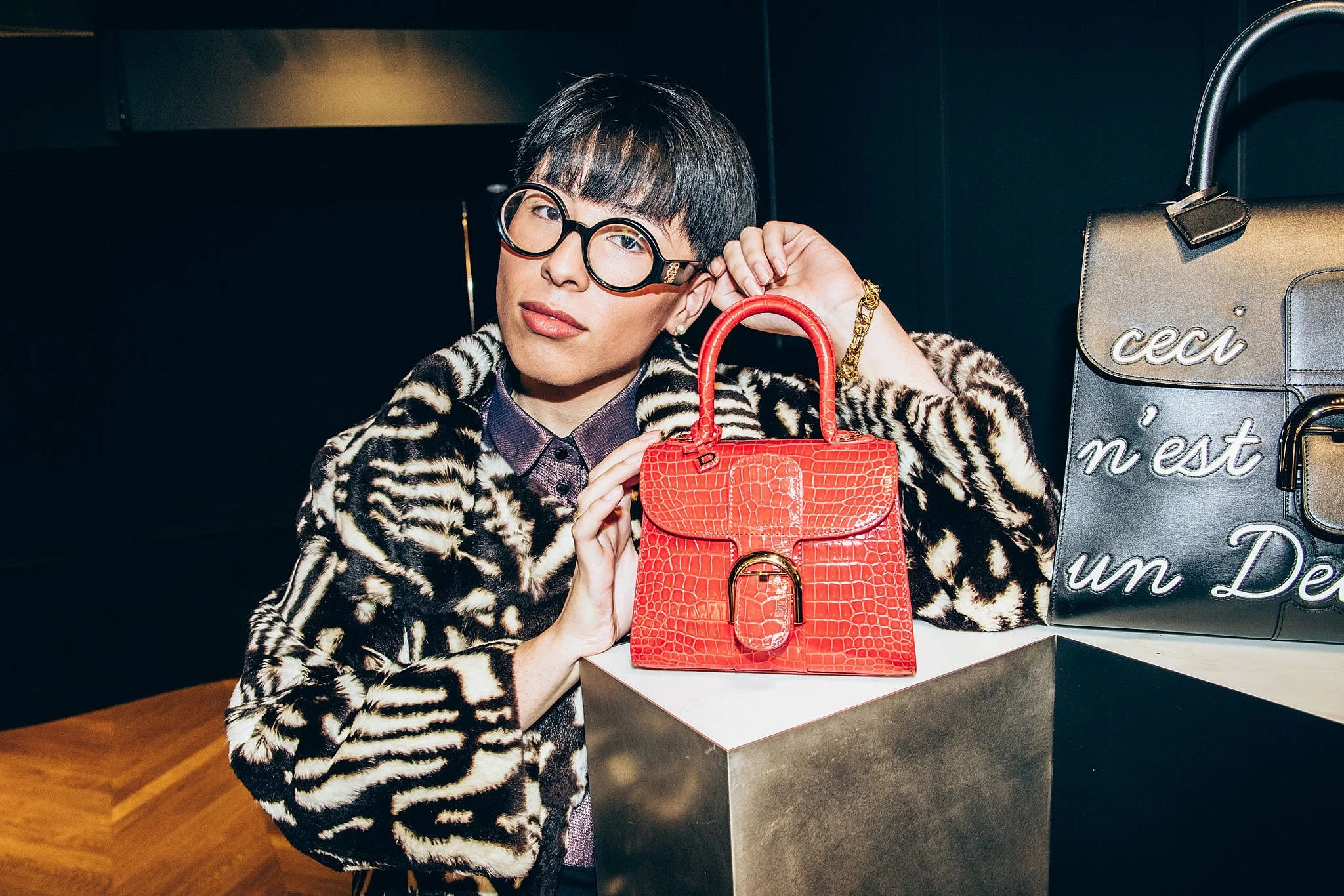 A woman with short dark hair, glasses, and a leopard print coat, sitting behind a display of designer handbags, including a red crocodile-patterned purse in the foreground and a black purse with white script on the right.