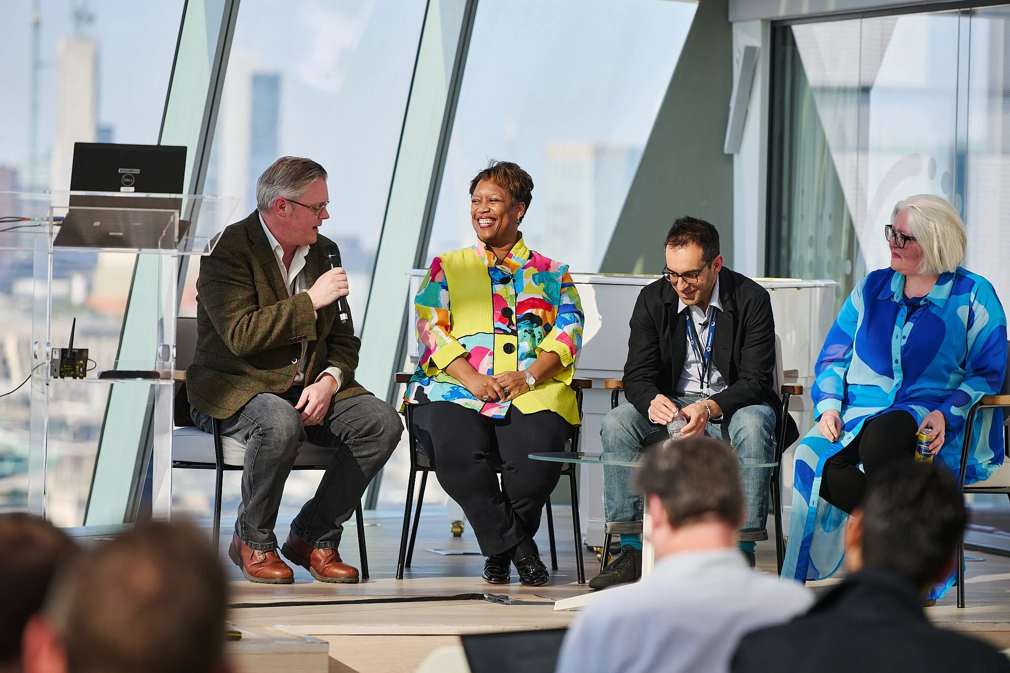 Four diverse individuals sitting on a stage in front of large windows, participating in a panel discussion; one man is speaking into a microphone, the woman in colorful attire is smiling, and the others are listening, with cityscape visible through t