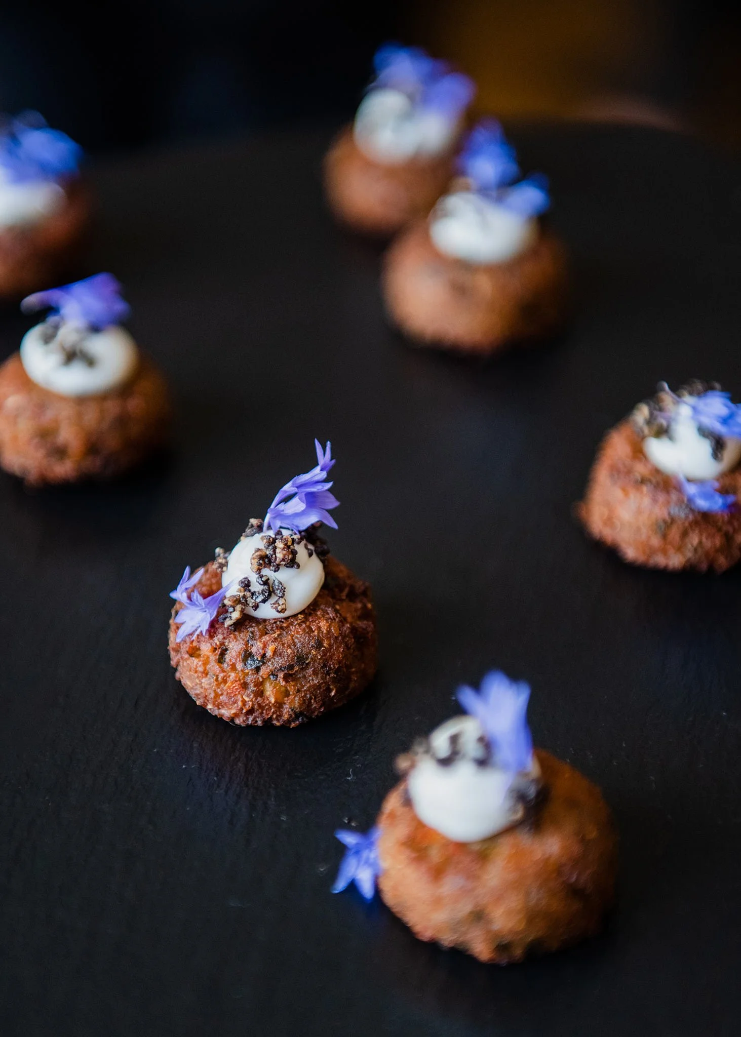 Crispy fried appetizers topped with white sauce and purple edible flowers on a black surface.