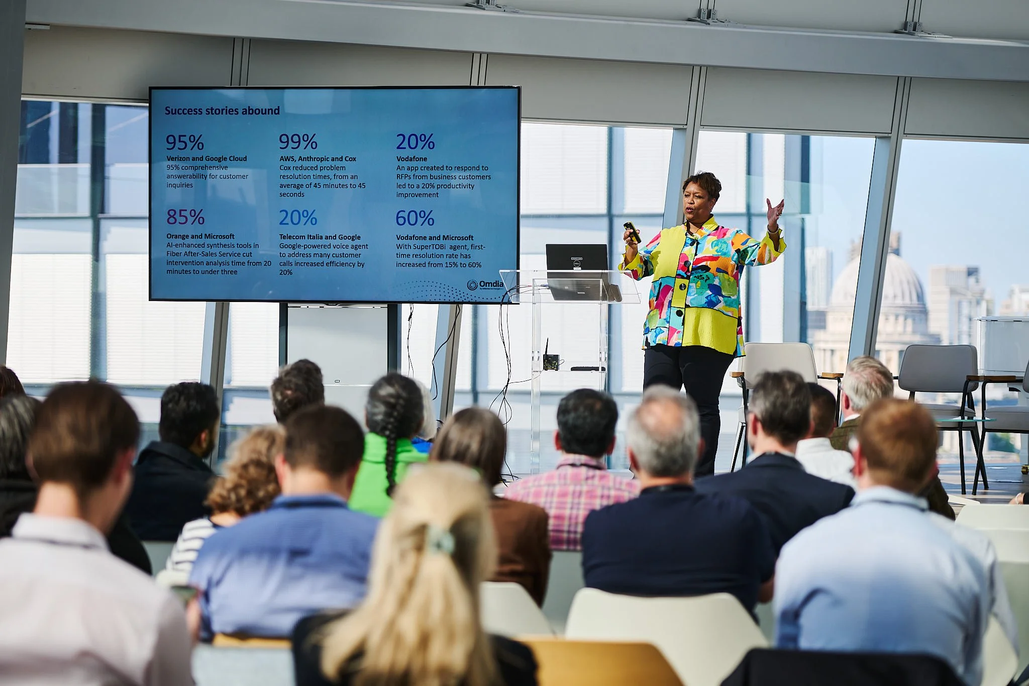 A woman giving a presentation in front of a large digital screen to an audience in a modern office space, with city buildings visible through large windows.