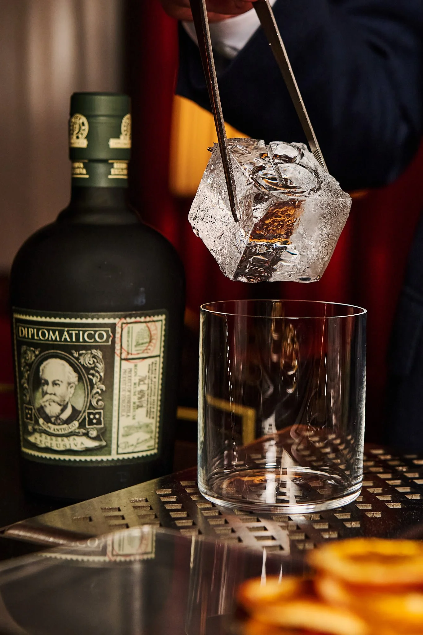 A bartender is preparing to pour a drink into a glass with a large ice cube using tongs, with a bottle of Diplomatico rum on the counter.